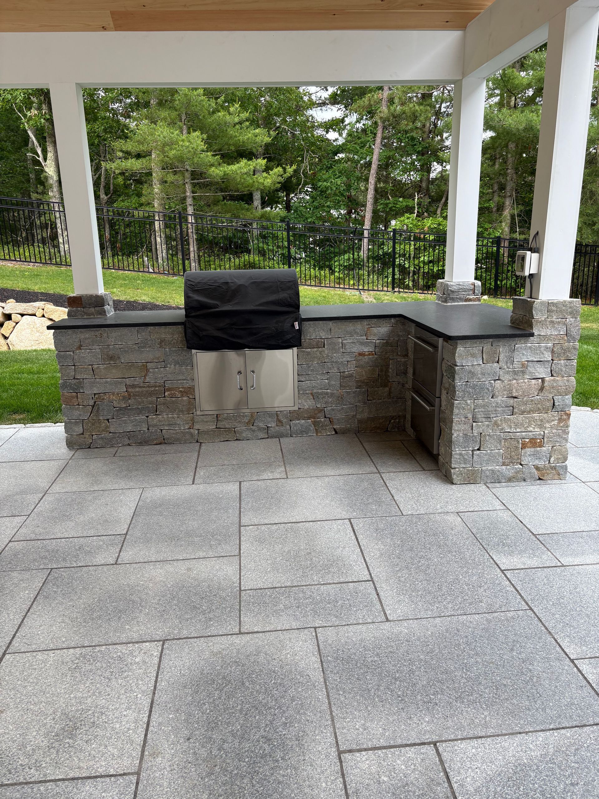 Covered outdoor stone grill with a black cover, set on a patio under a pavilion.