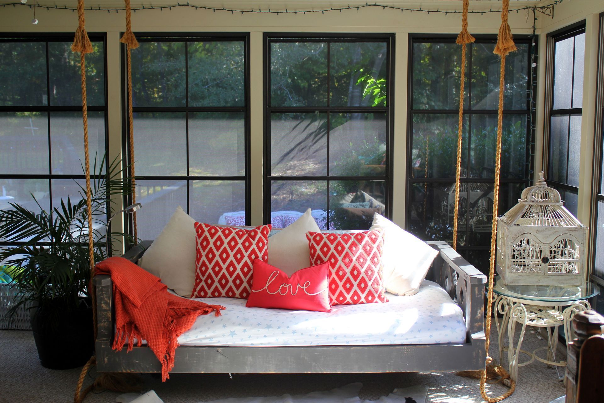 Cozy porch daybed with red pillows, orange throw, hanging swings, and plants in a sunlit screened room