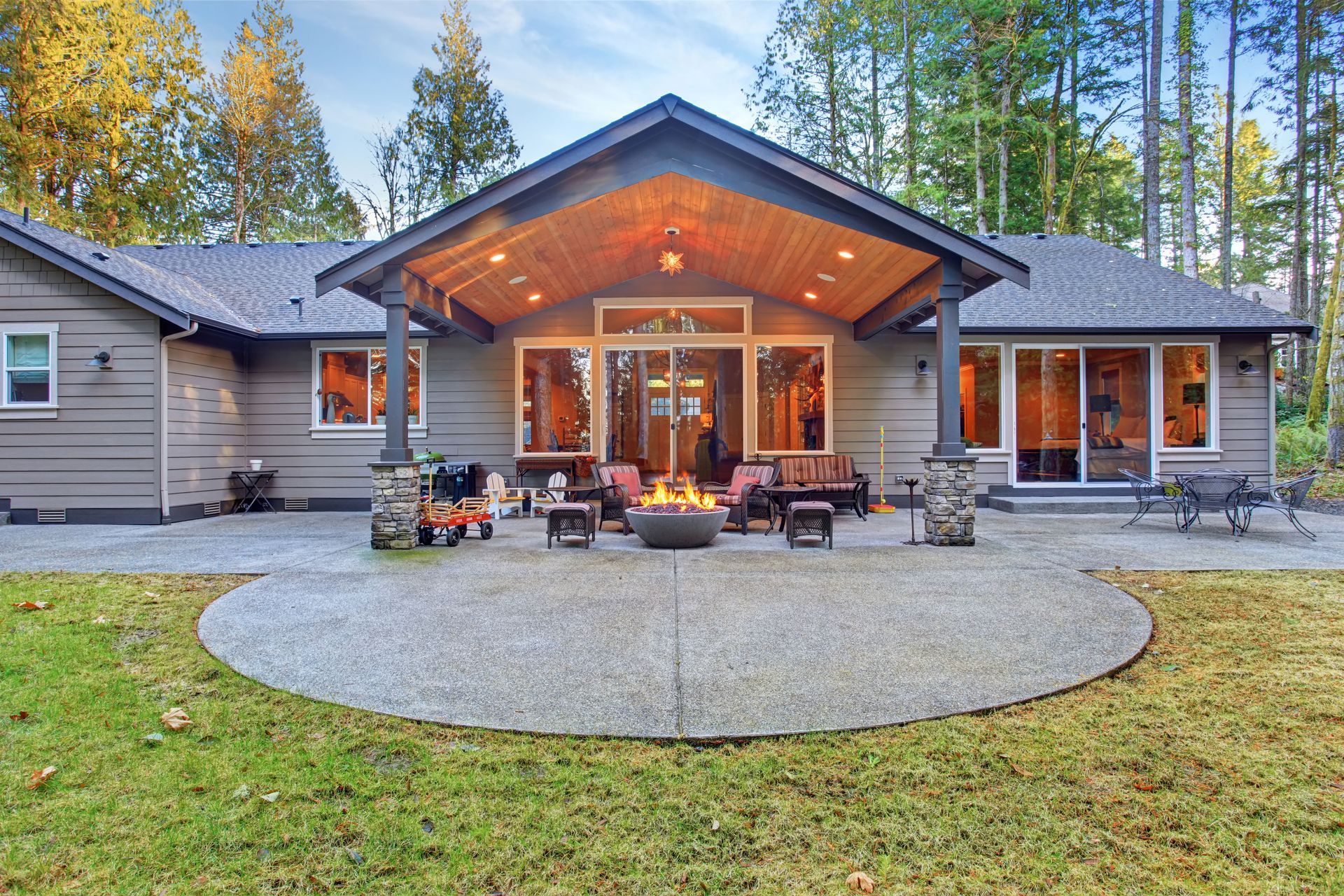Modern house with a large patio, outdoor seating, and a fire pit in a wooded yard at dusk