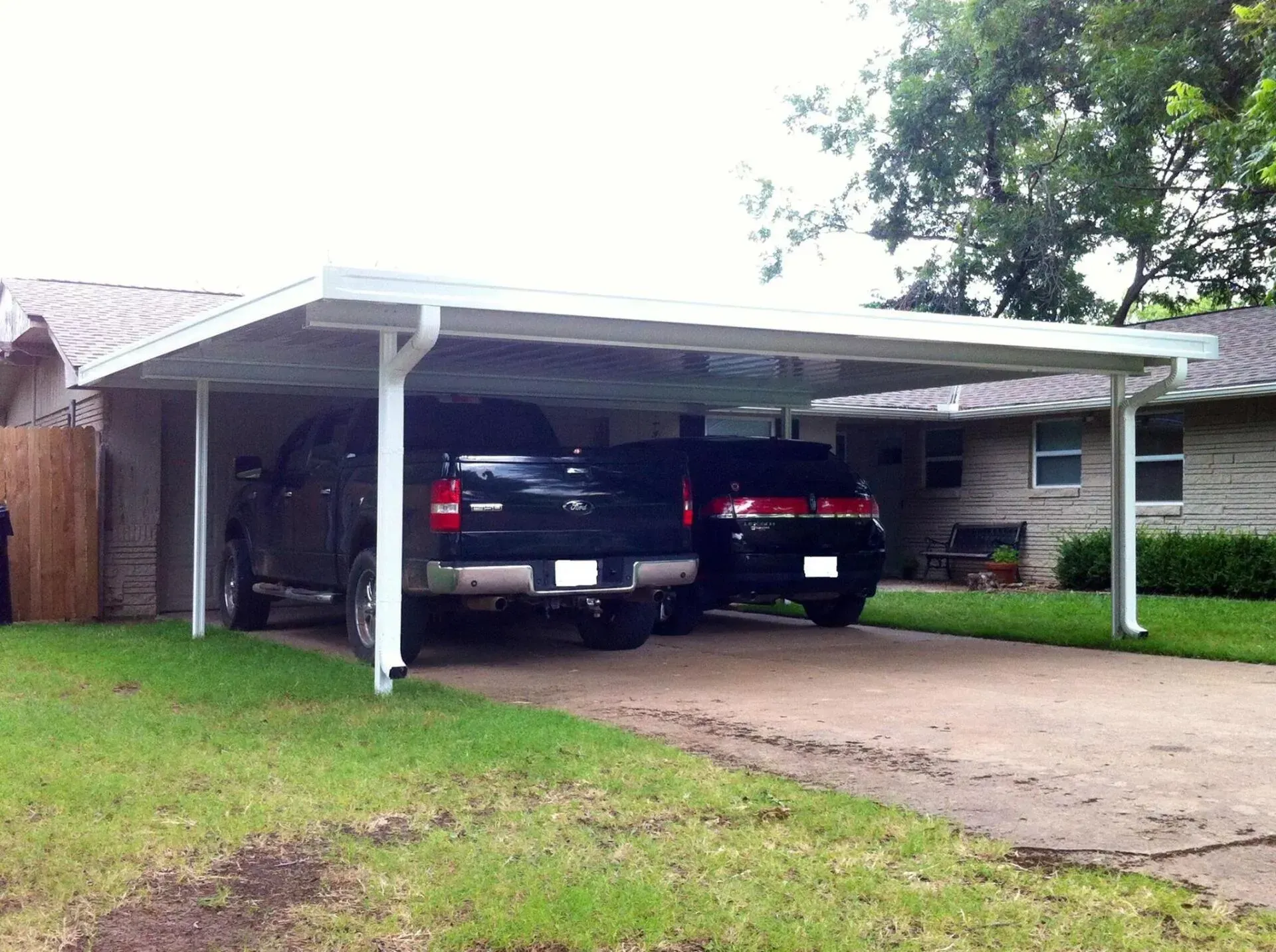 Two cars parked under a white carport beside a house with a grassy yard.