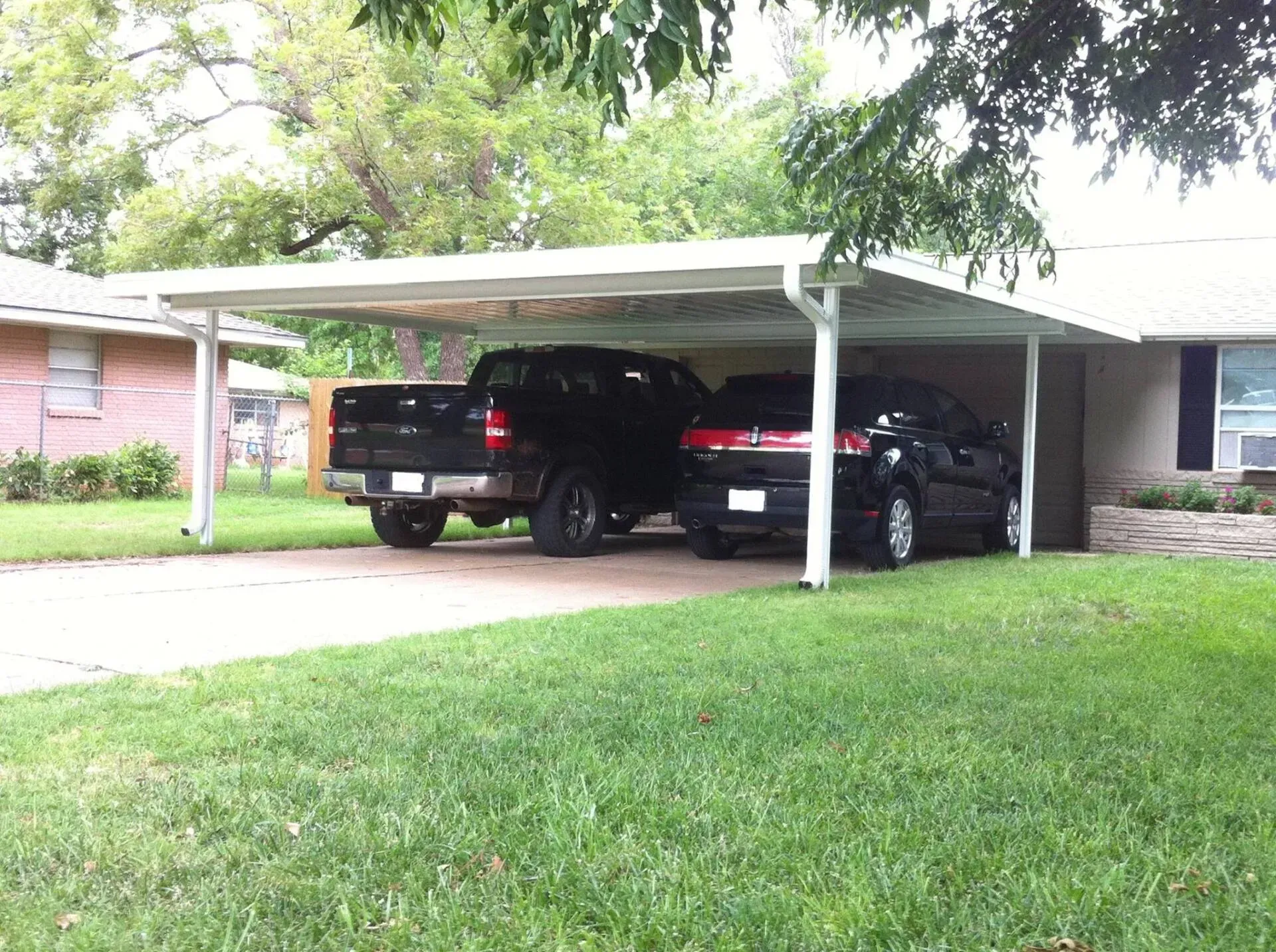 Two cars parked under a carport beside a brick house and green lawn