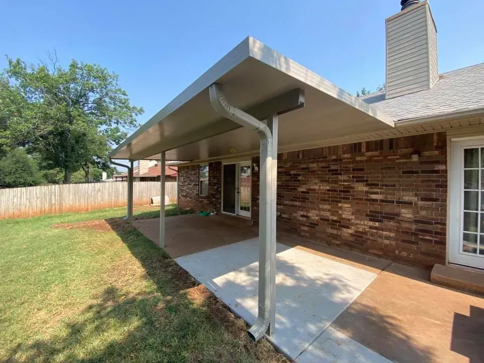 Covered concrete patio beside brick house with metal awning and backyard fence