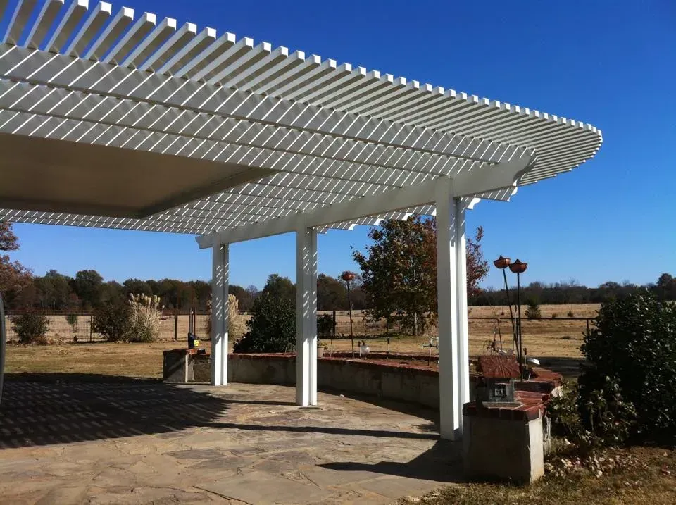 White pergola over a stone patio with planters, set in a sunny open field under a blue sky