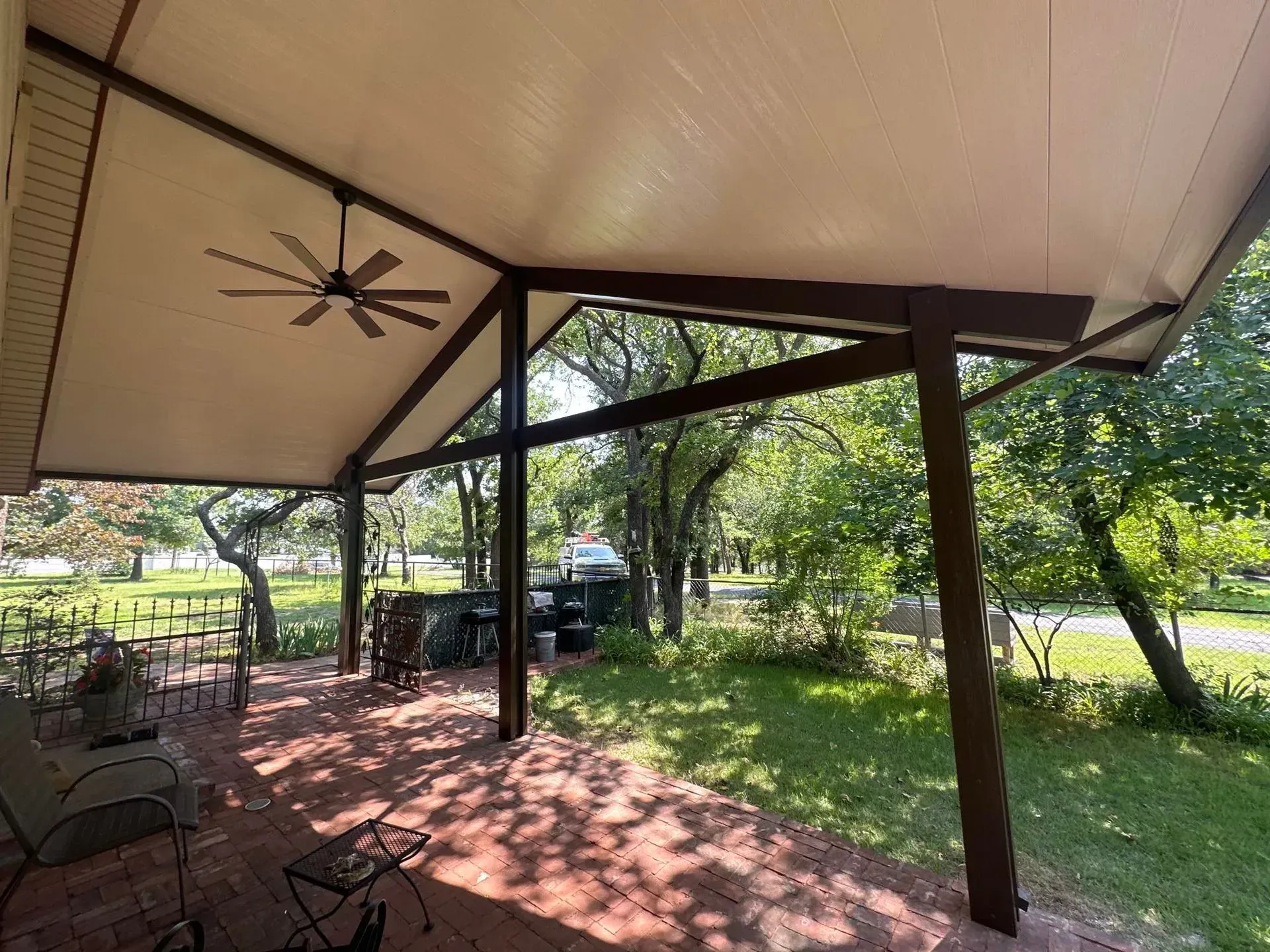 Shaded patio with ceiling fan and metal chairs overlooking a sunny grassy yard and trees