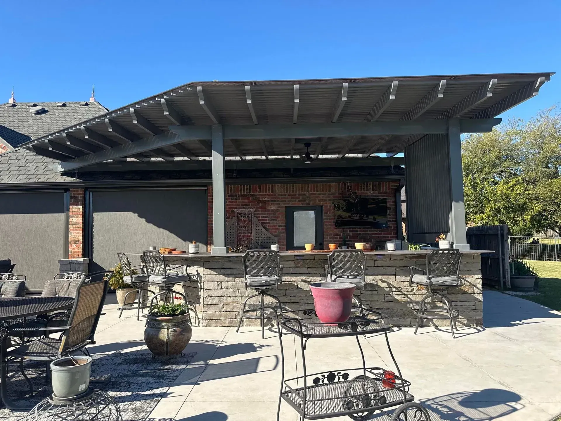 Outdoor patio with tables and chairs under a covered pergola, with potted plants and a grill in front.
