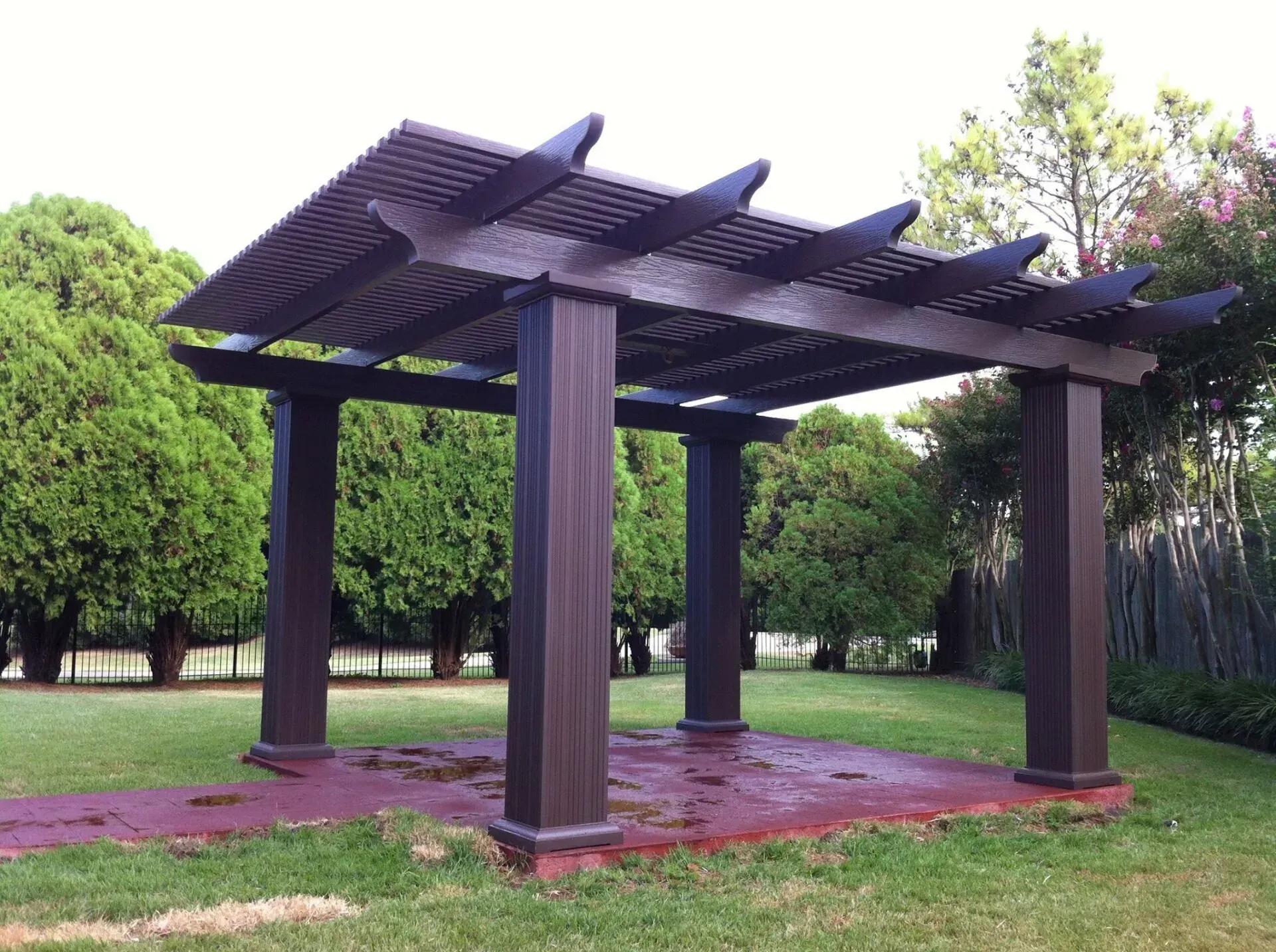 Wooden pergola gazebo with a slatted roof in a green park, surrounded by trees and grass.
