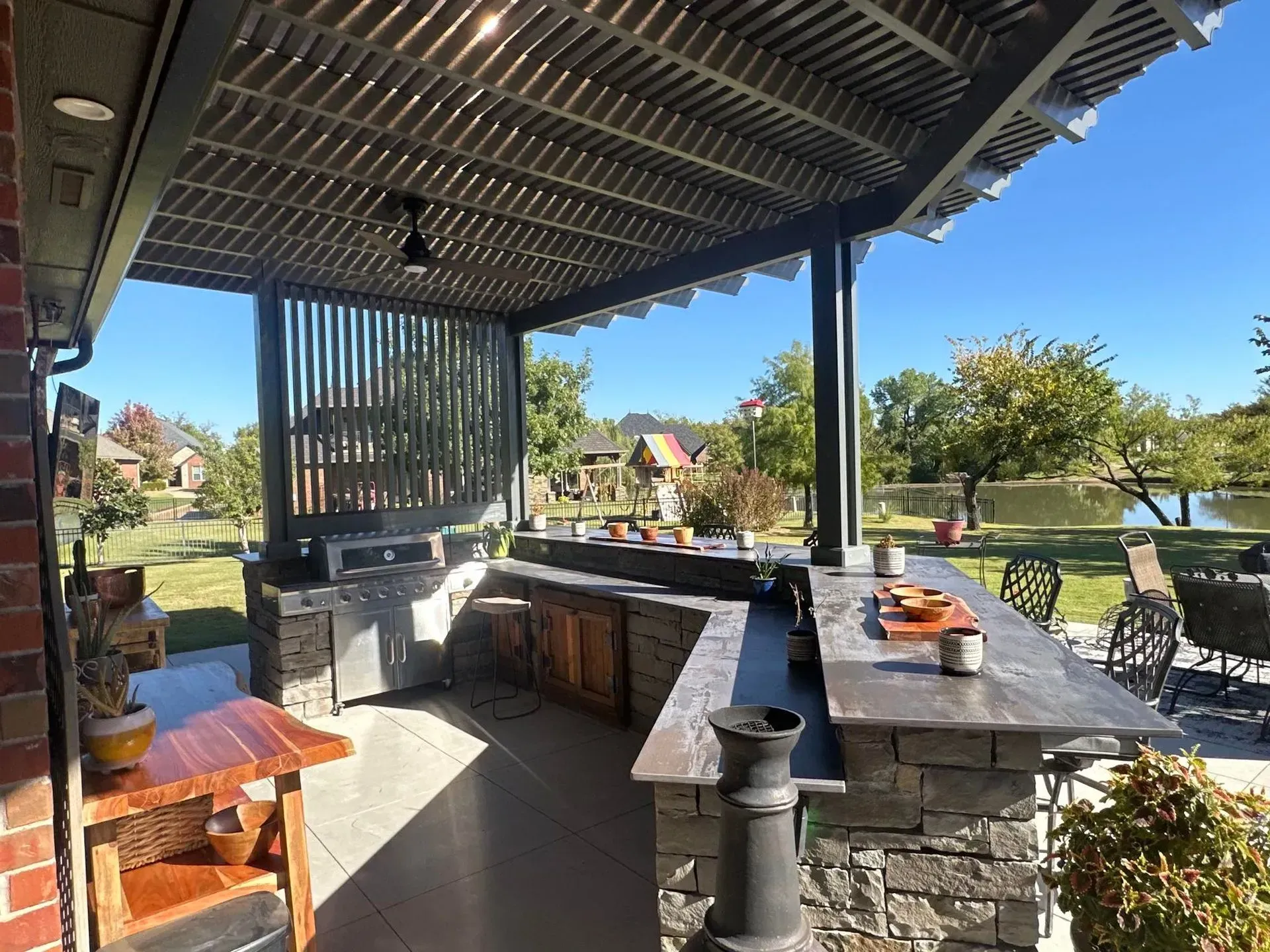 Covered outdoor bar and patio with stone counter, wooden stools, and a sunny garden view.