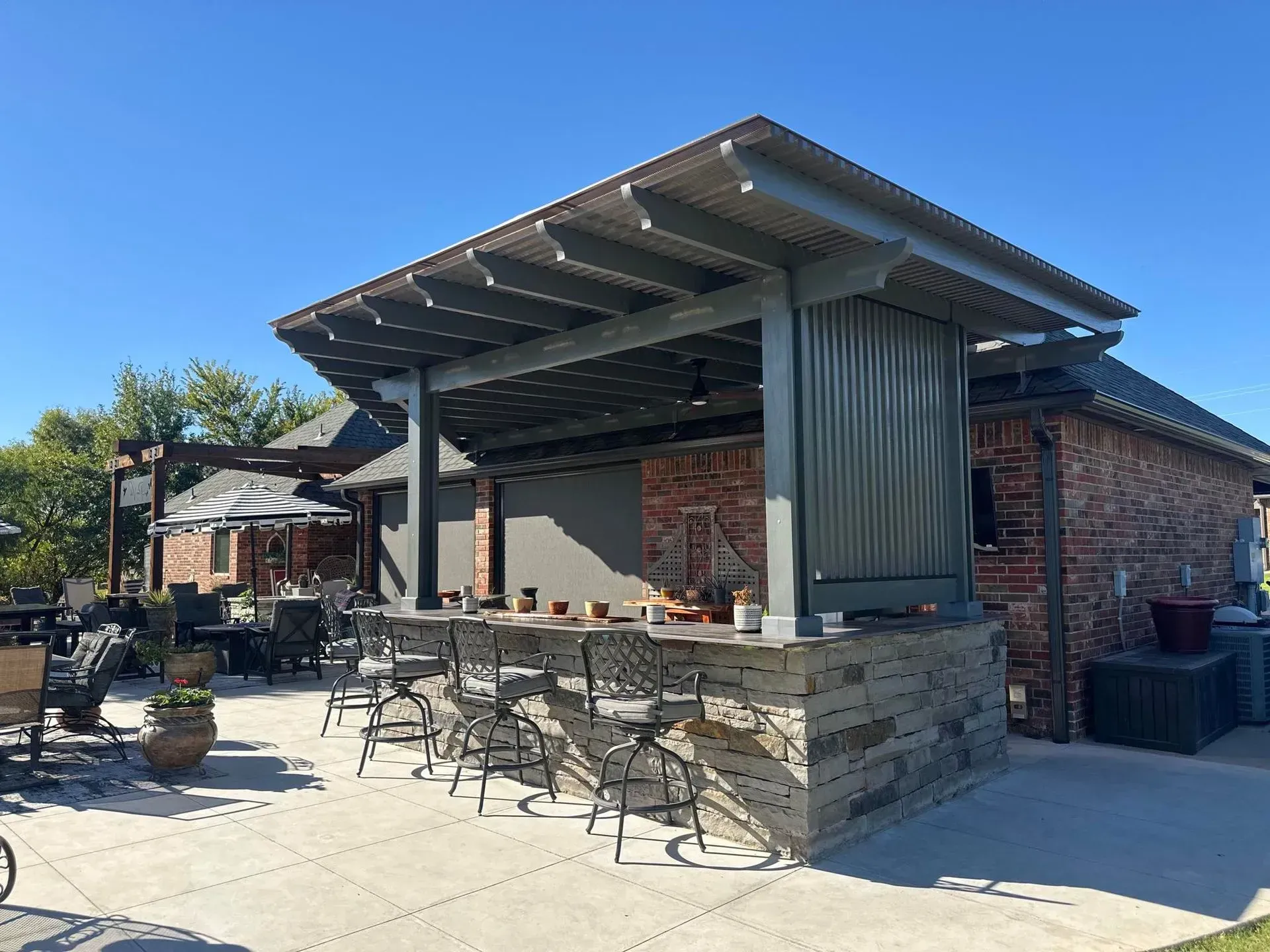 Outdoor restaurant patio with stone bar, metal pergola roof, and empty tables and chairs in sunlight