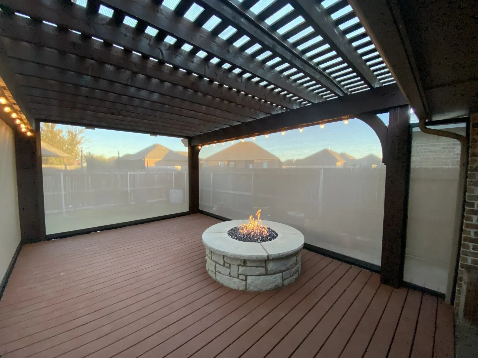 Covered deck with a round stone fire pit and brown wood flooring, enclosed by mesh screens.
