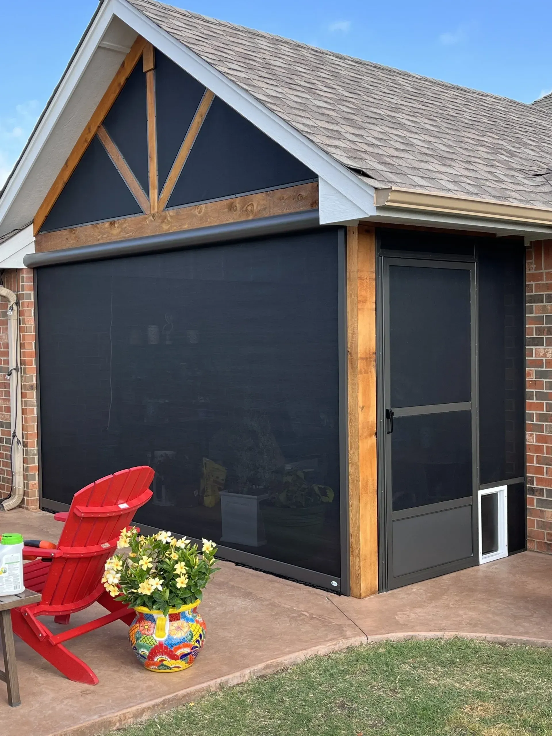 Black screened porch/sunroom on a house with red chairs and potted flowers on the patio.