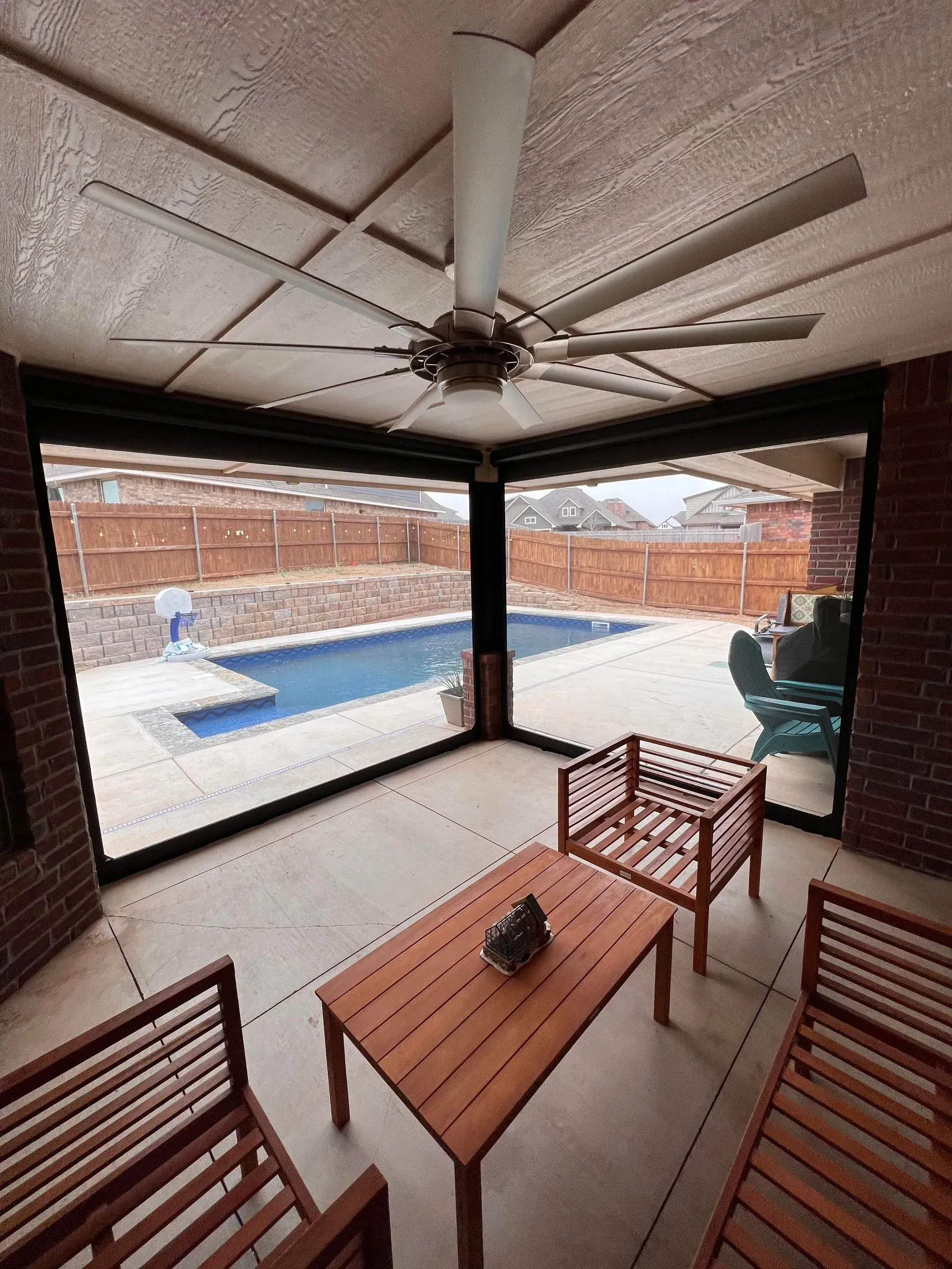 Covered patio with ceiling fan, wooden table and chairs, overlooking a fenced backyard pool