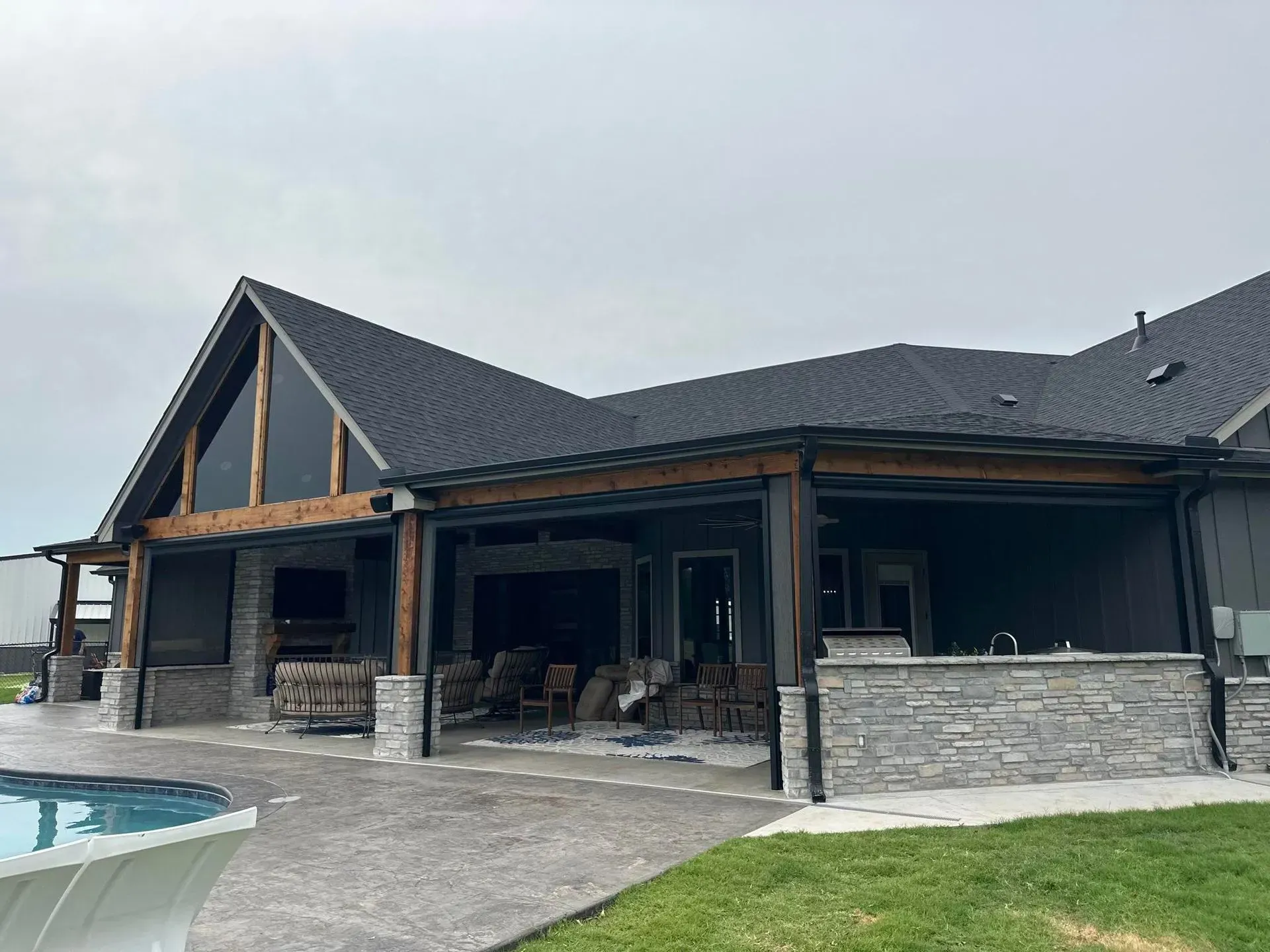 Modern stone house with a dark roof and covered patio beside a pool and lawn
