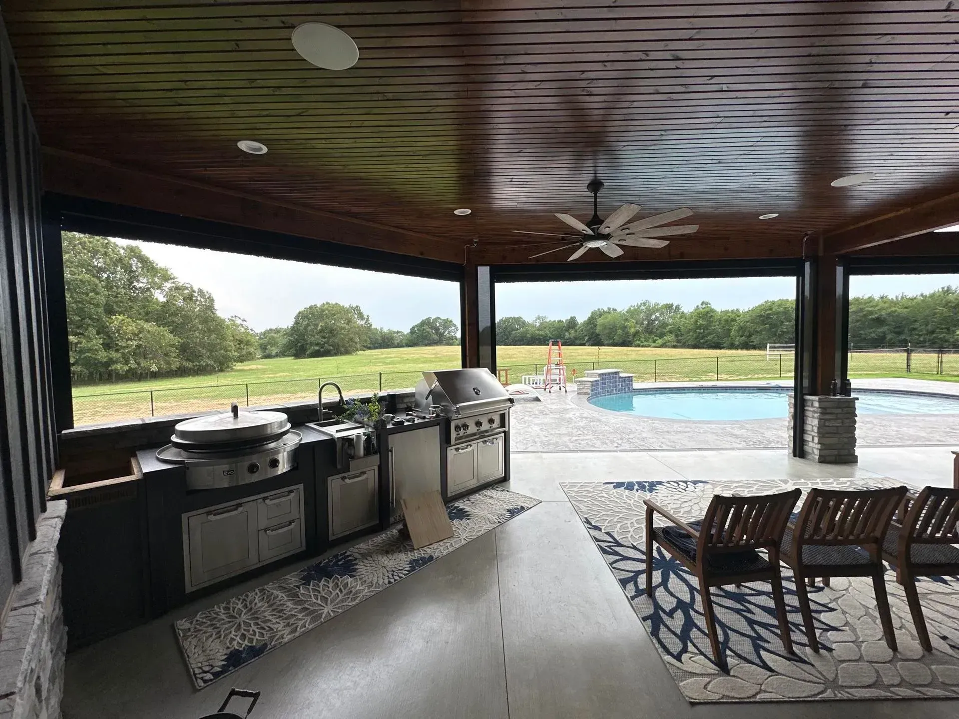 Covered patio with grill and dining table overlooking a pool and green landscape.