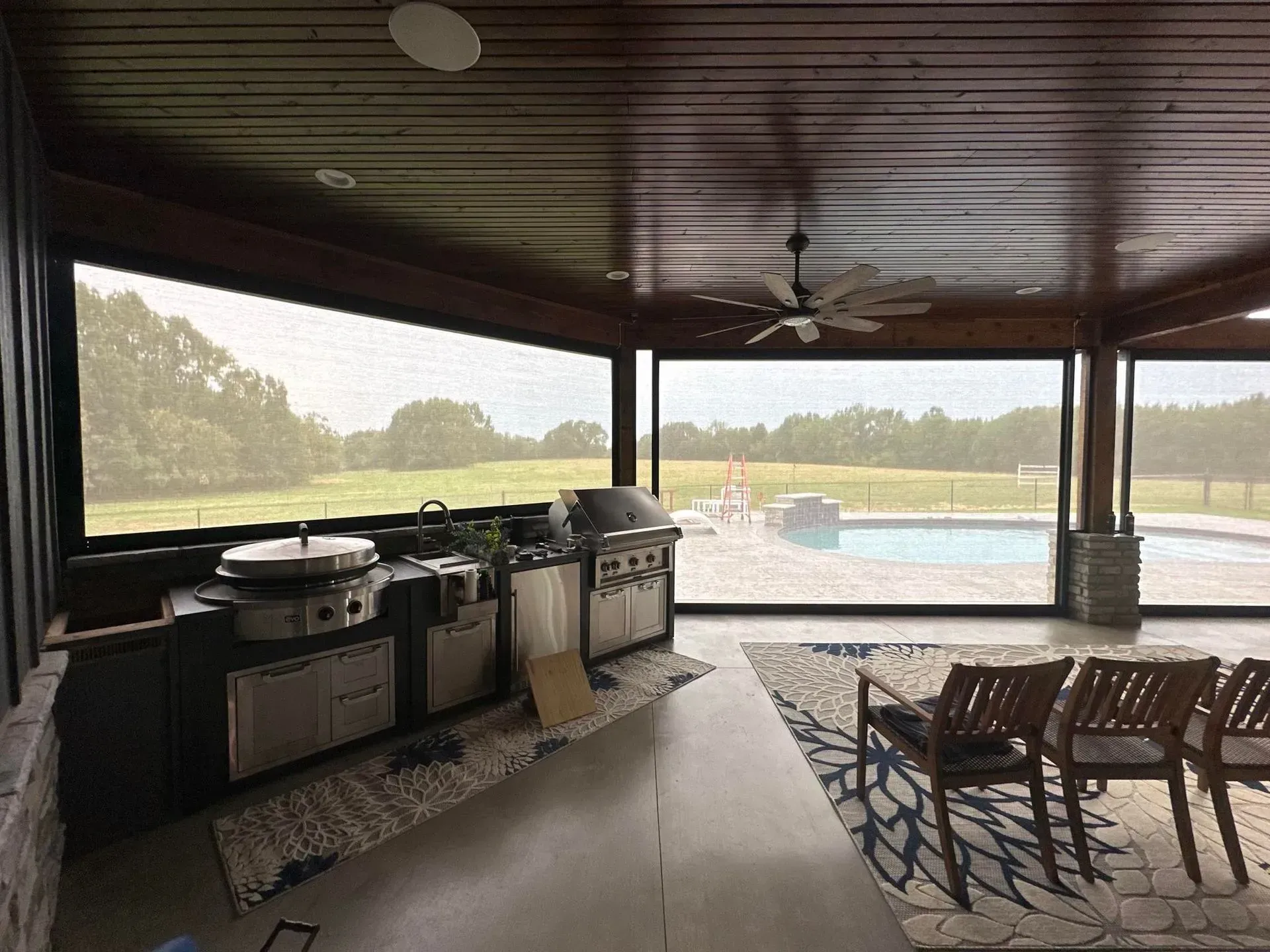 Covered patio with outdoor kitchen, seating, and a view of a pool and open field beyond.