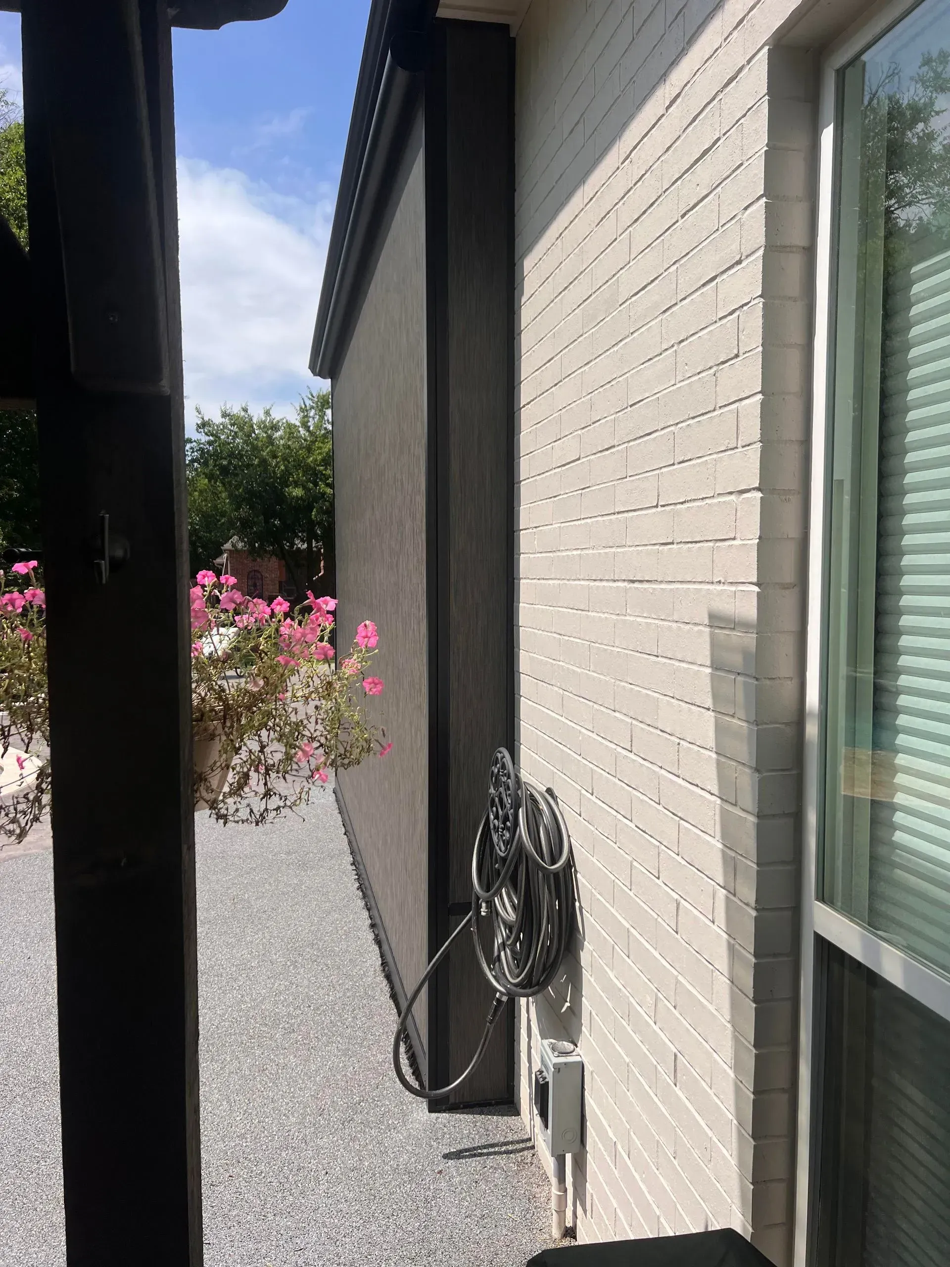 Narrow side yard beside a white brick house, with a coiled hose and pink flowers in the distance.