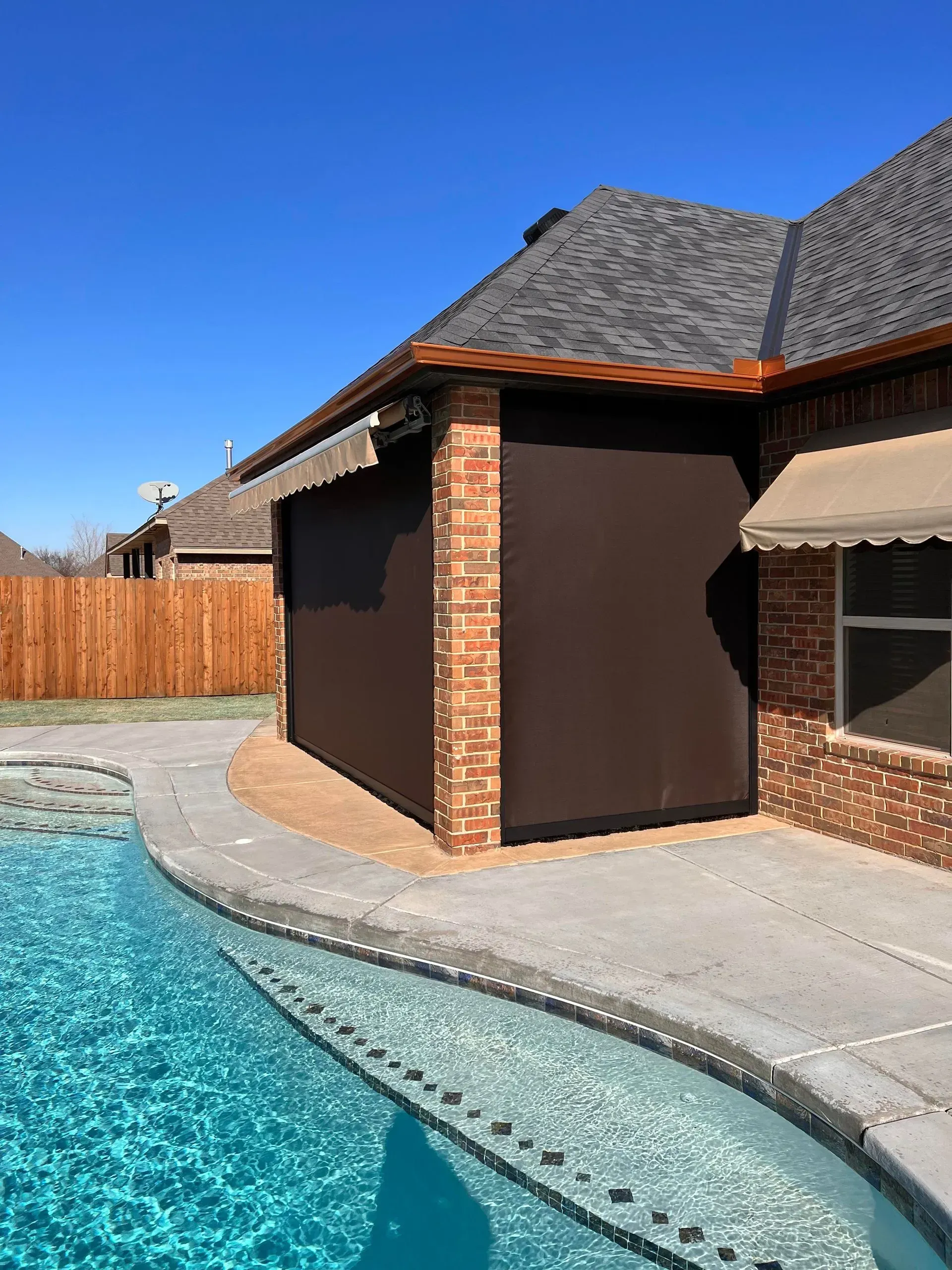 Backyard pool beside a brick house with a gray roof and wooden fence under a clear blue sky