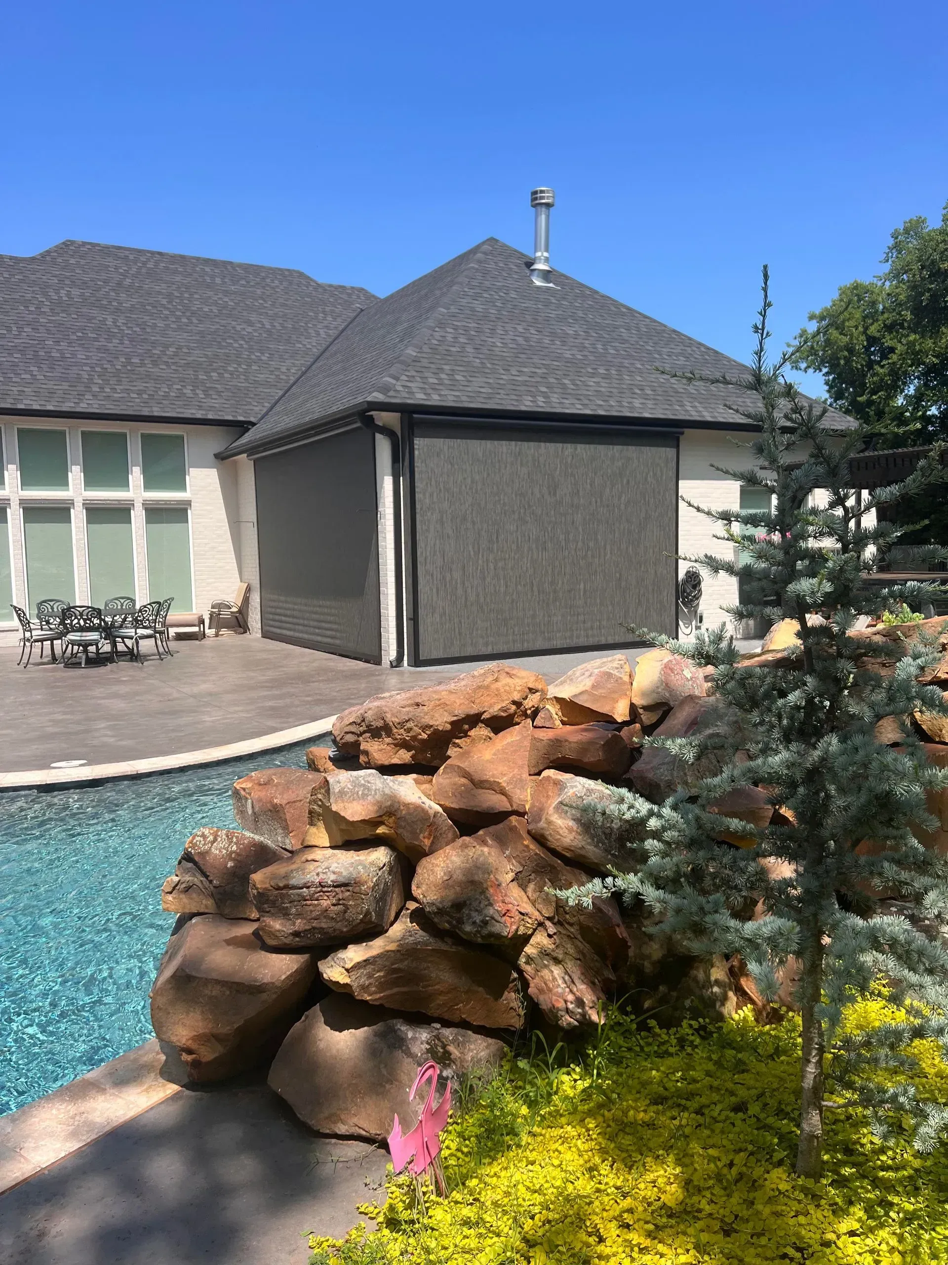 Backyard pool with stacked rocks, flowering shrubs, and a house under a blue sky