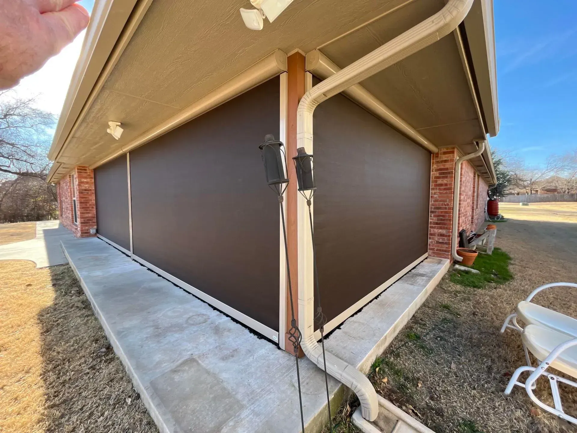 Closed brown patio shades on the corner of a house with a concrete walkway and lawn.