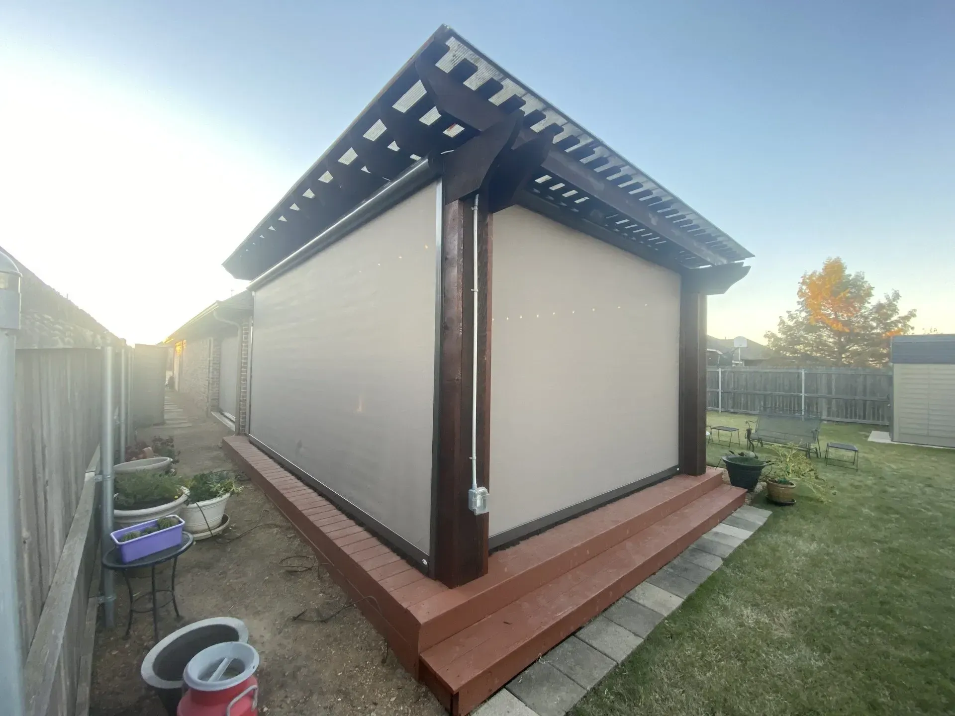 Small backyard shed with beige walls, dark roof, and wooden deck beside a fenced lawn.