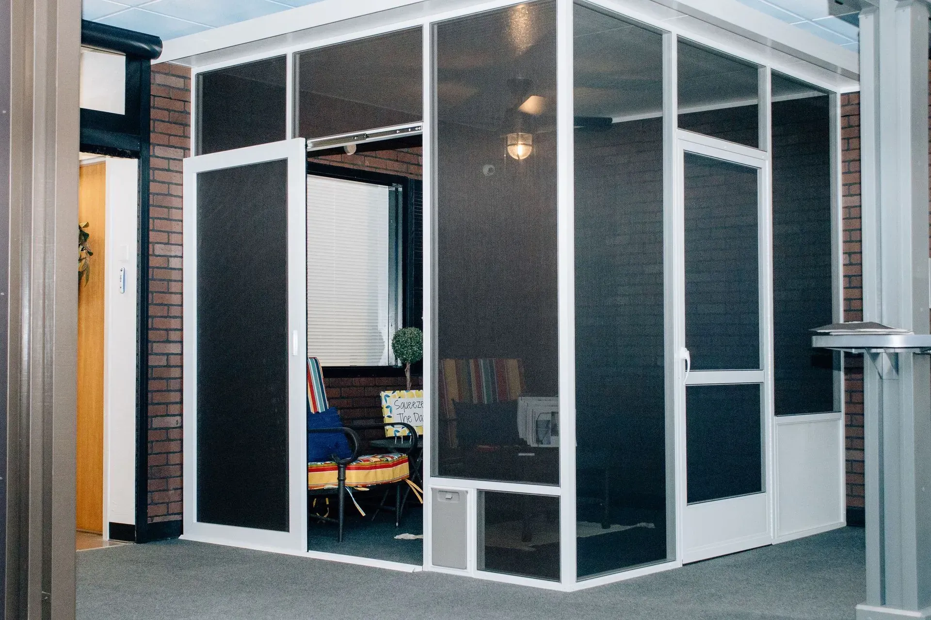 Modern glass-walled office booth with open door and chairs inside, in a carpeted indoor space