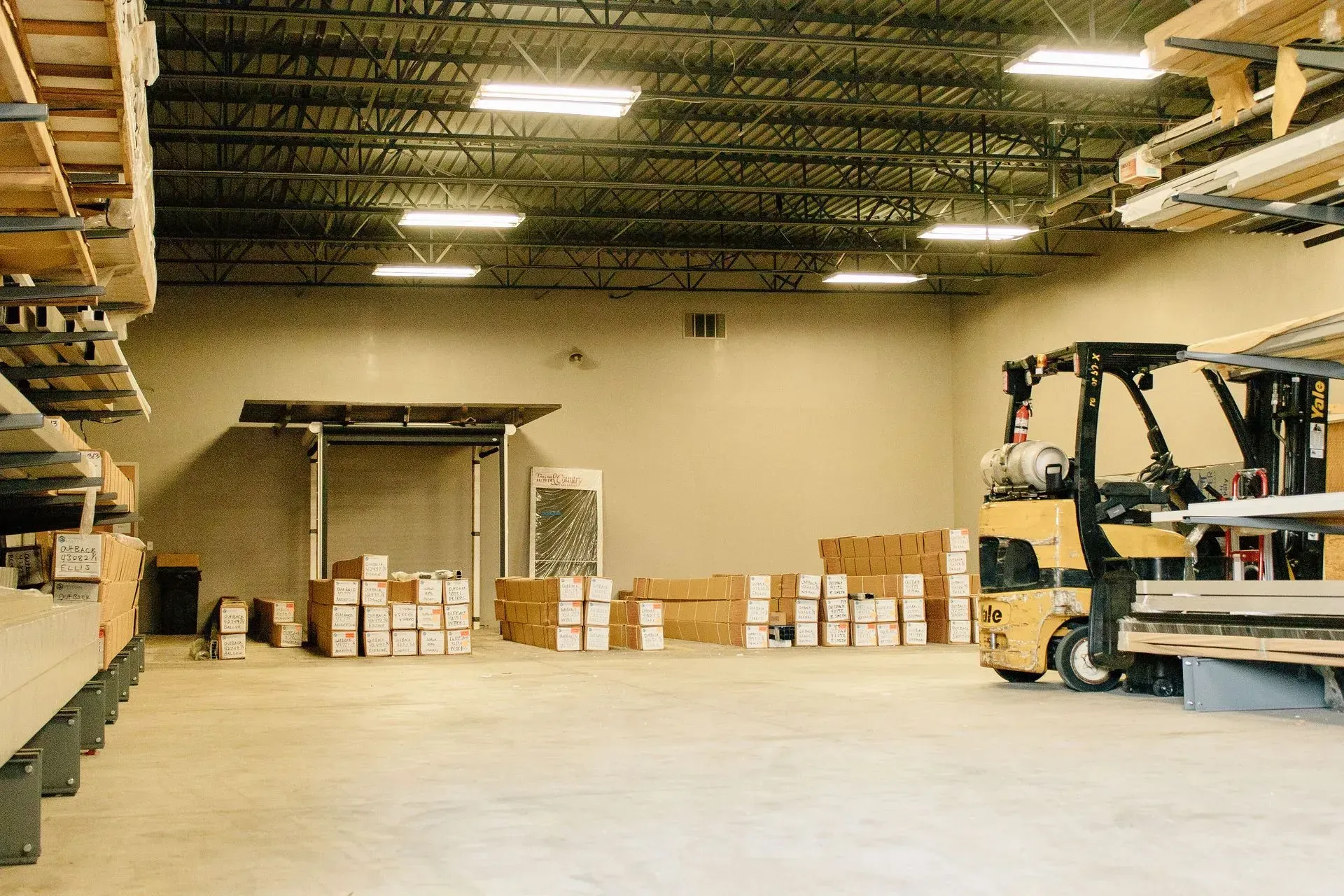 Warehouse interior with stacked boxes, shelving, and a forklift under bright overhead lights
