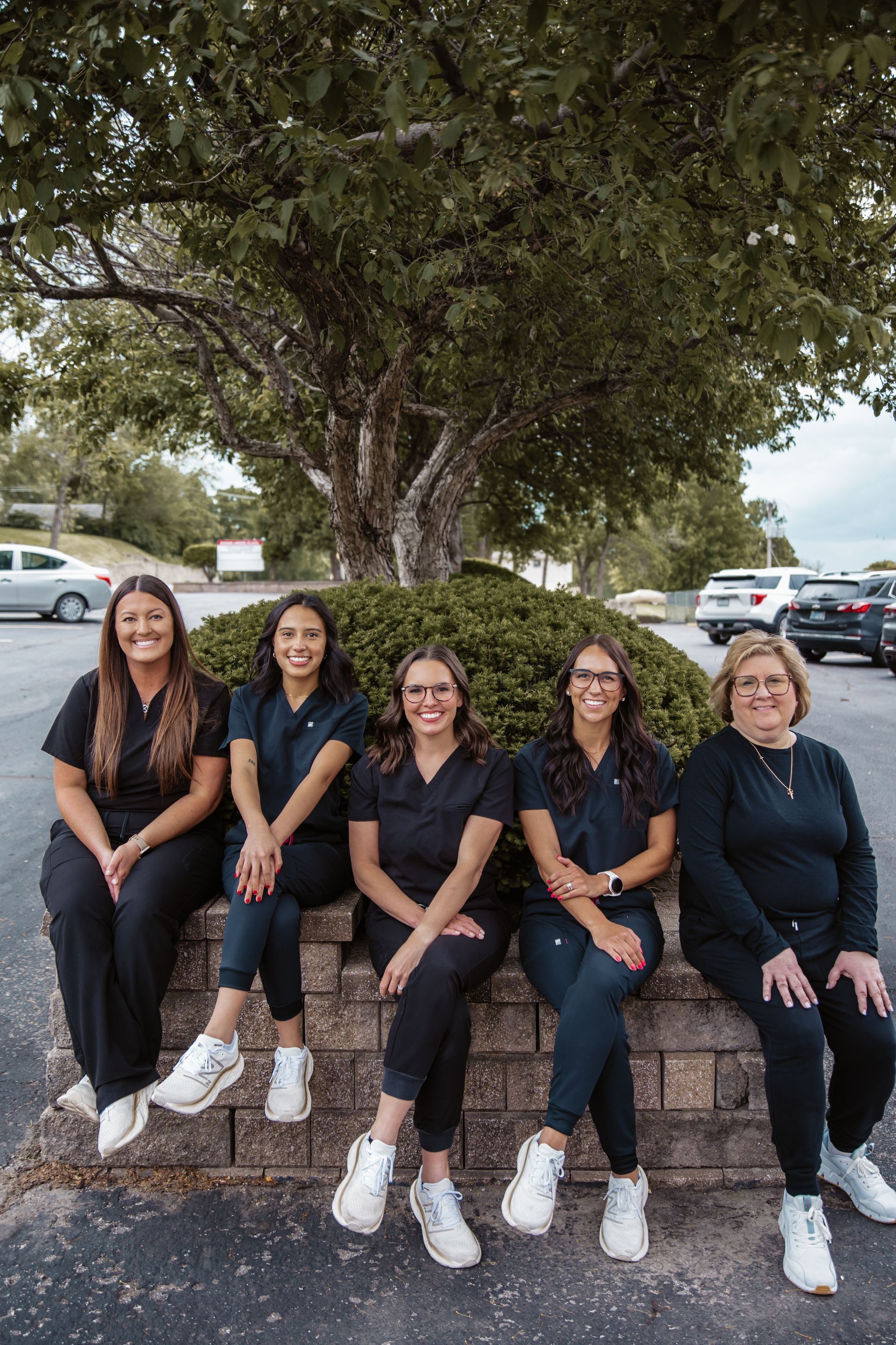 Five people in black scrubs, sitting on a brick wall outdoors, smiling at the camera.