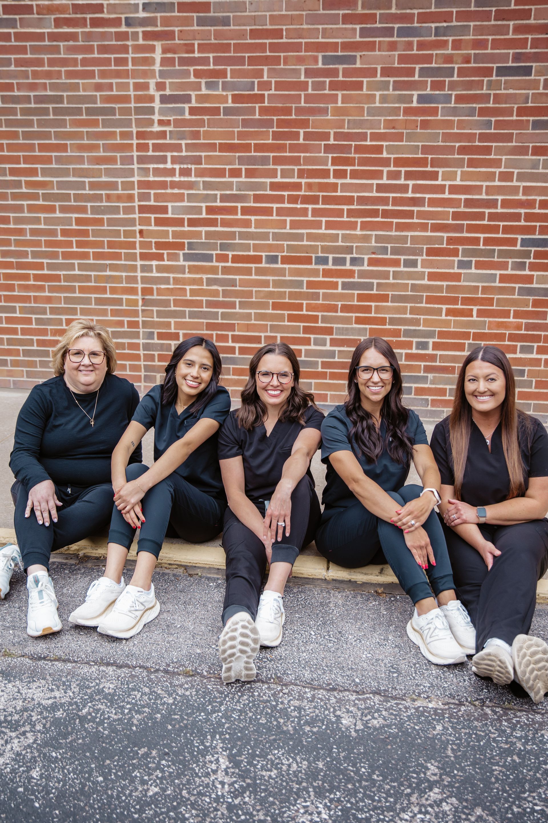 Five women in black scrubs sit in front of a brick wall, smiling.