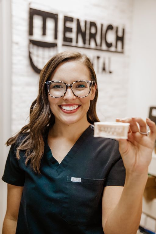 Woman in scrubs and glasses smiles, holding dental mold in front of 