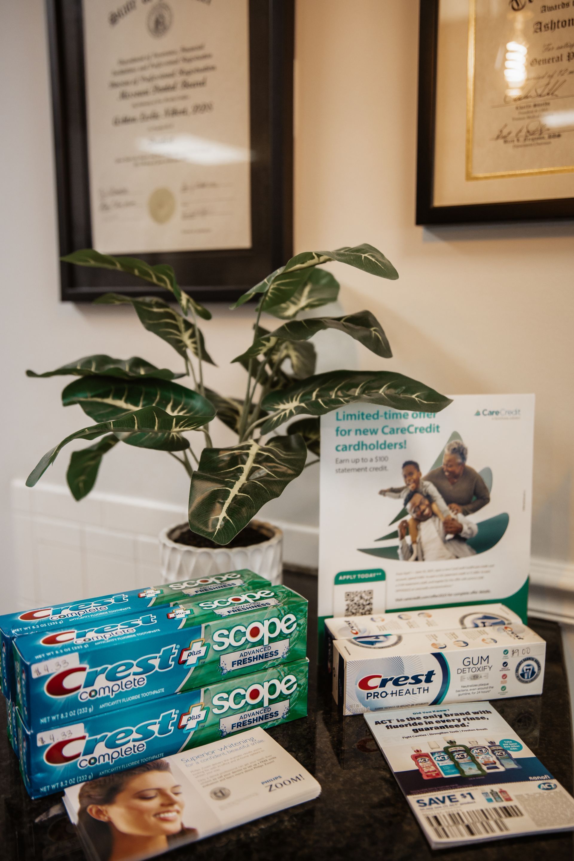 Toothpaste, brochures, and a plant on a desk under framed documents.