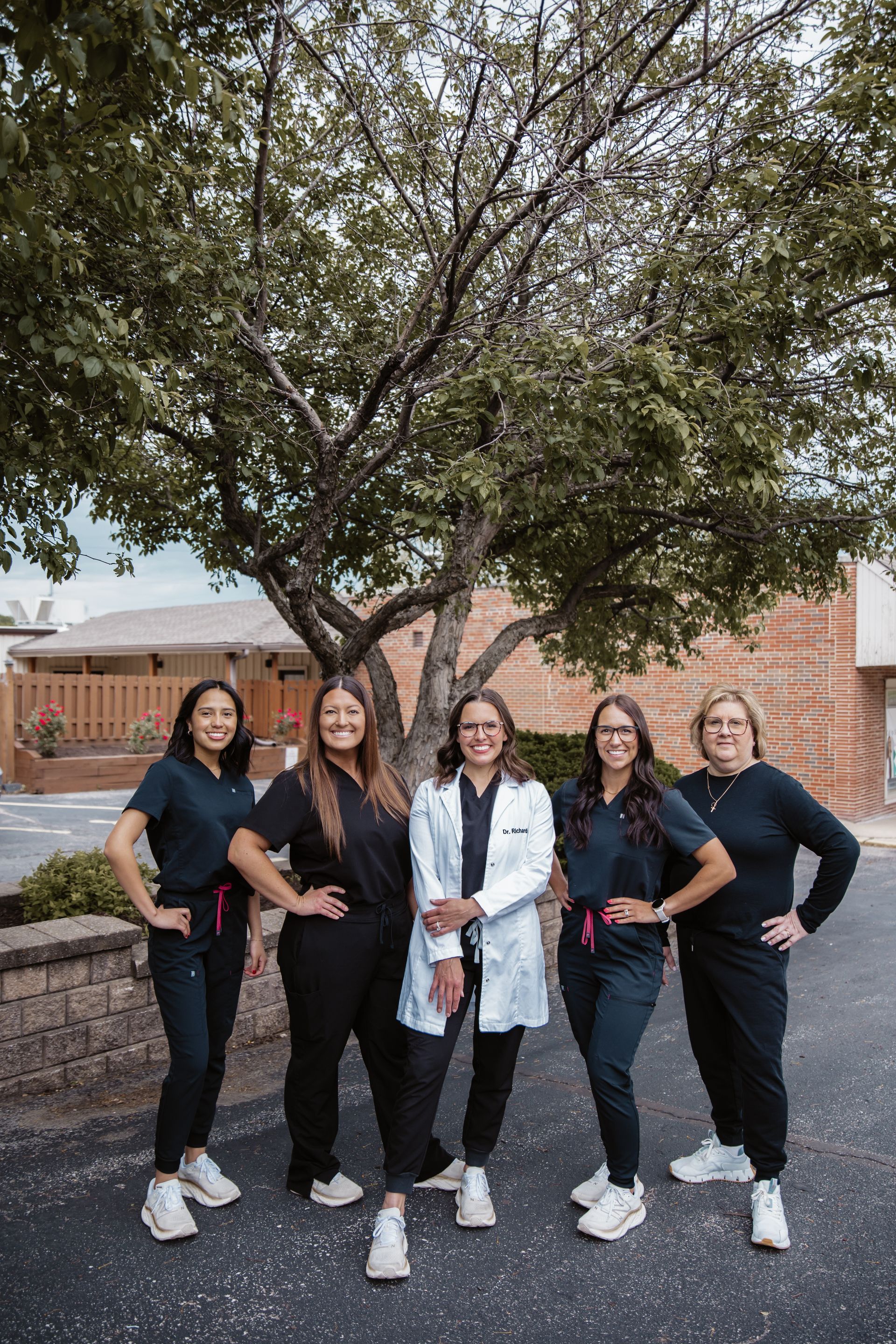 Five people wearing scrubs pose in front of a building with a tree.