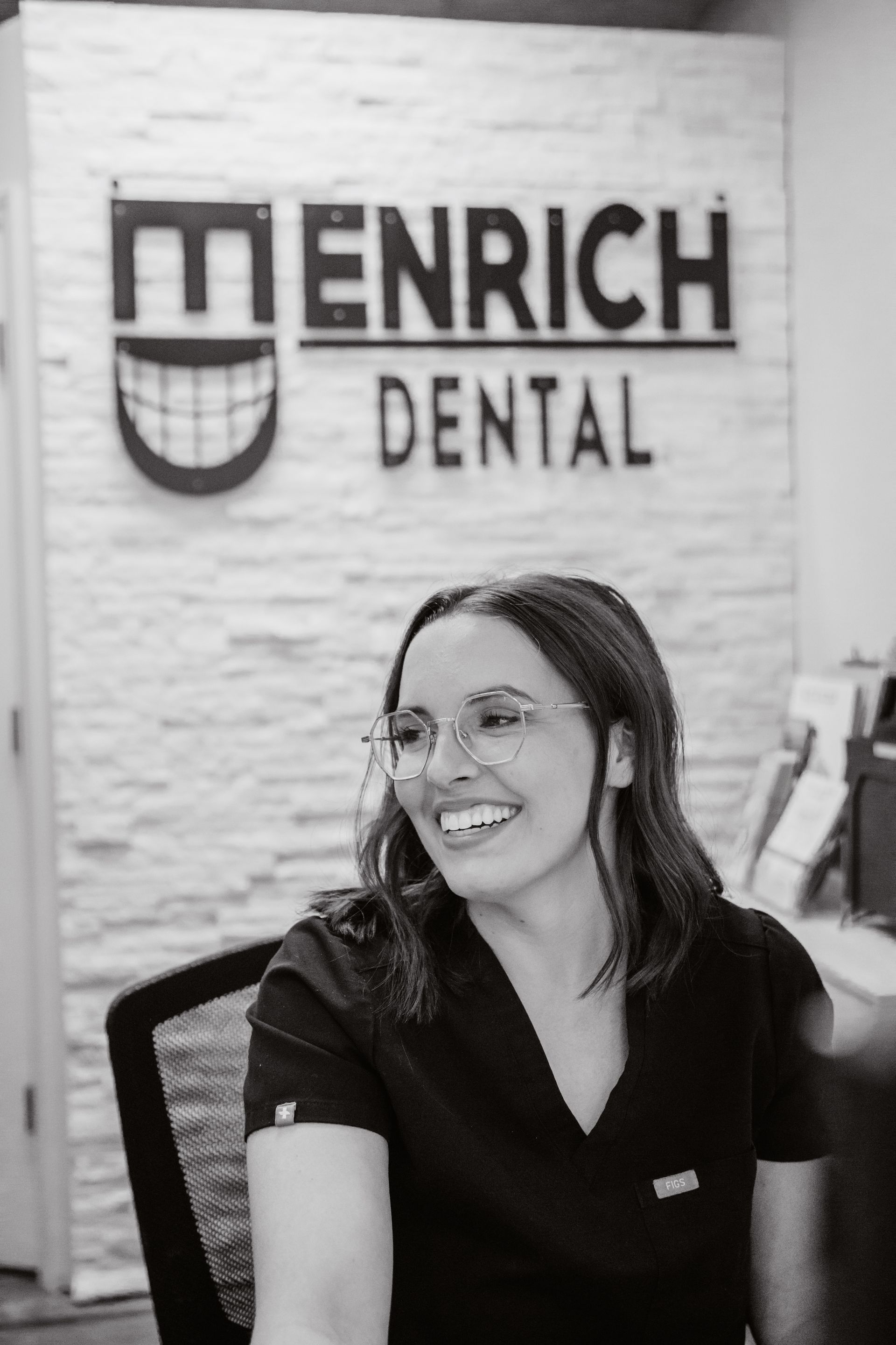 Smiling person in scrubs and glasses, seated near 
