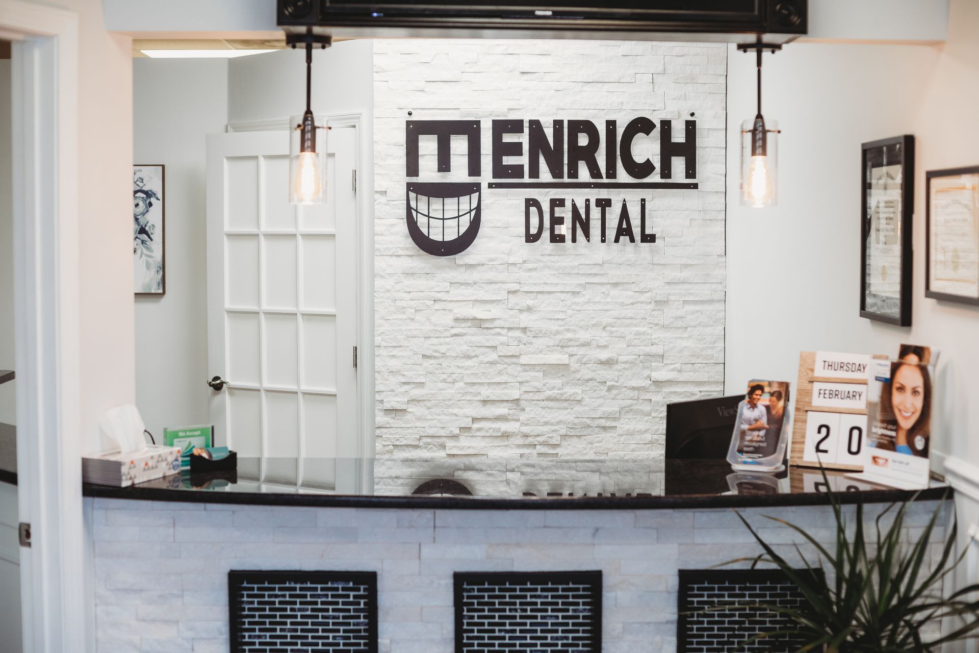 Reception area of Enrich Dental; white brick wall with logo, black counter, door on left, two hanging lights.