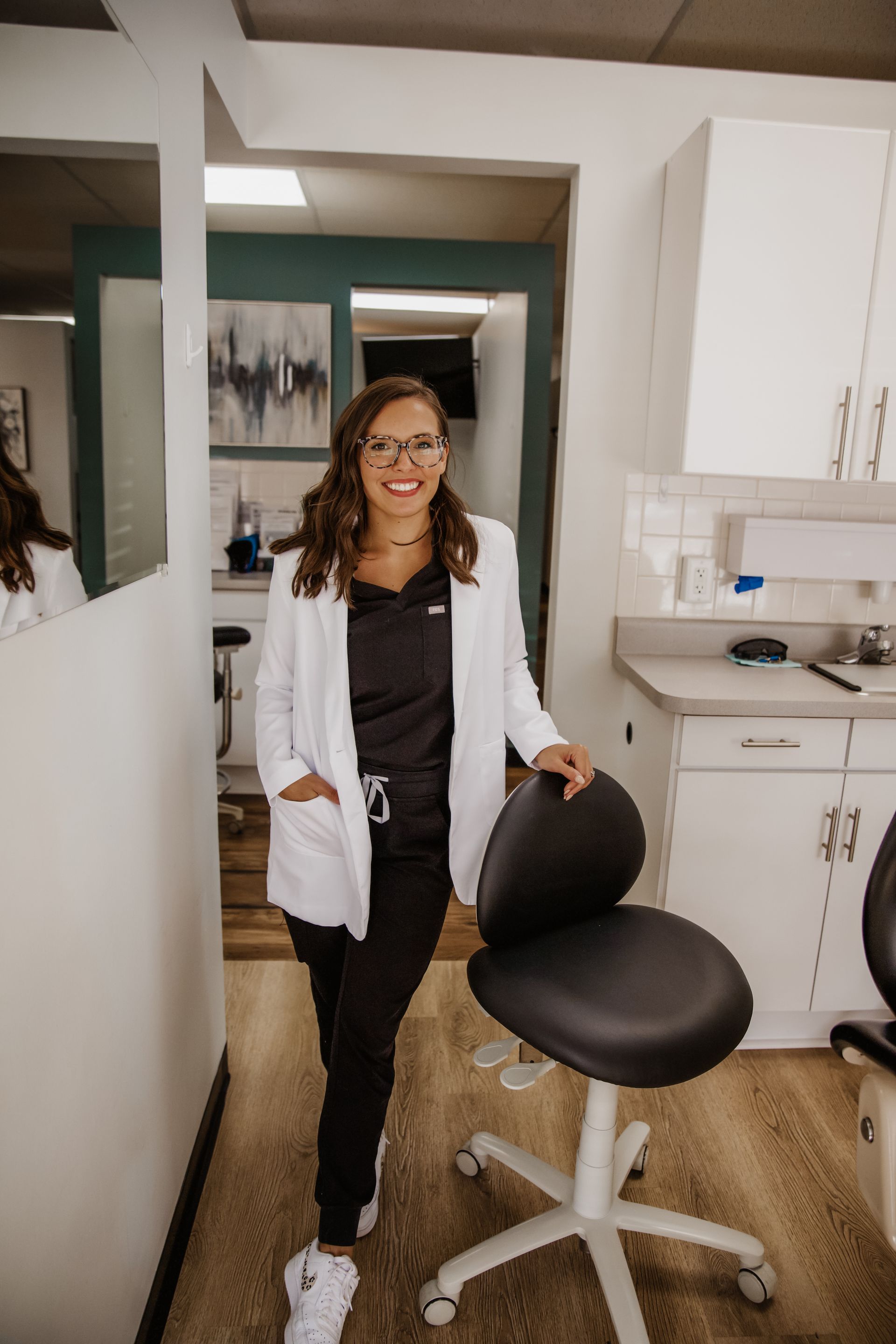 Woman in white lab coat smiles in a dental office, leaning on a black chair.