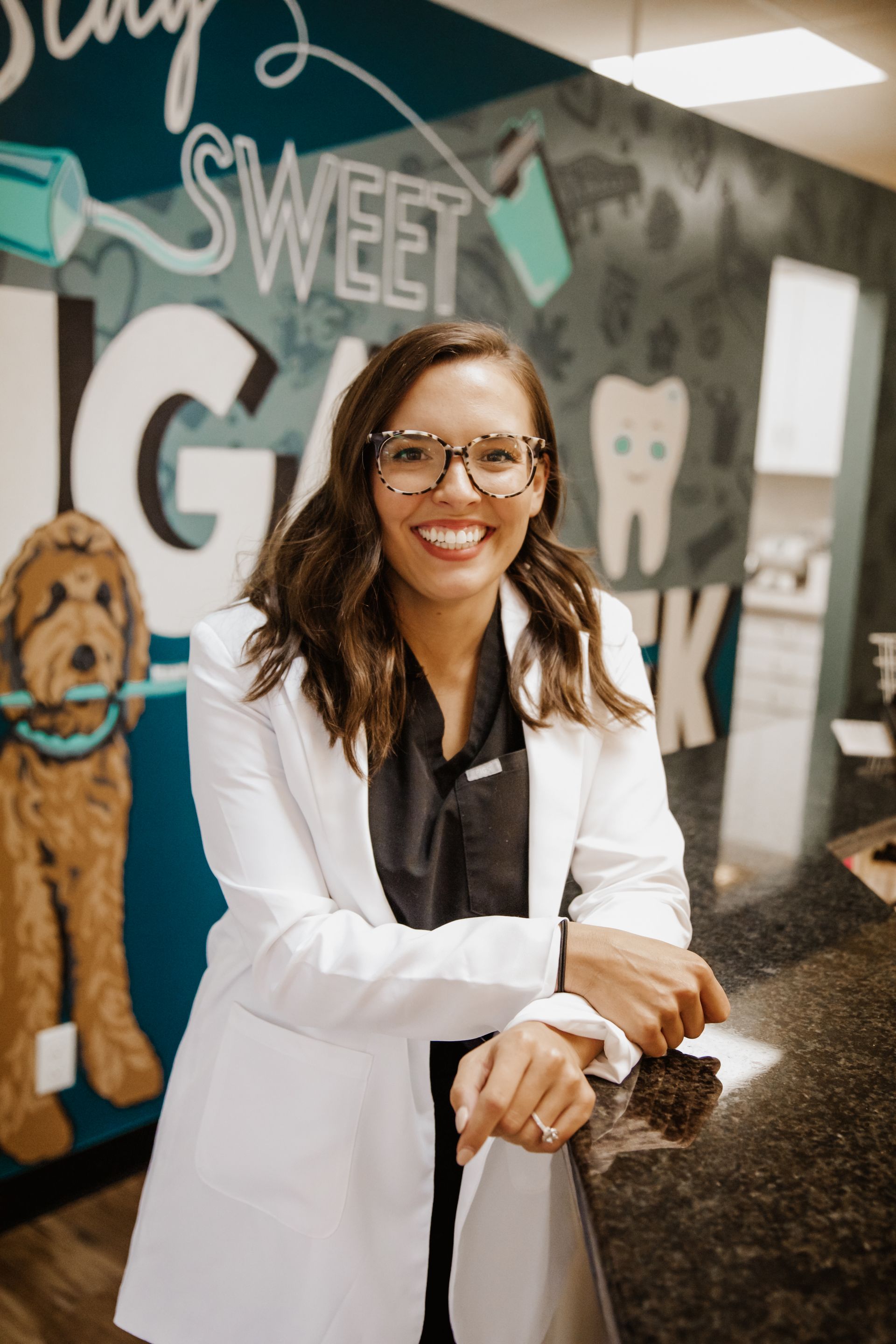 Woman in white lab coat smiles near a counter, dental office setting, background decor.