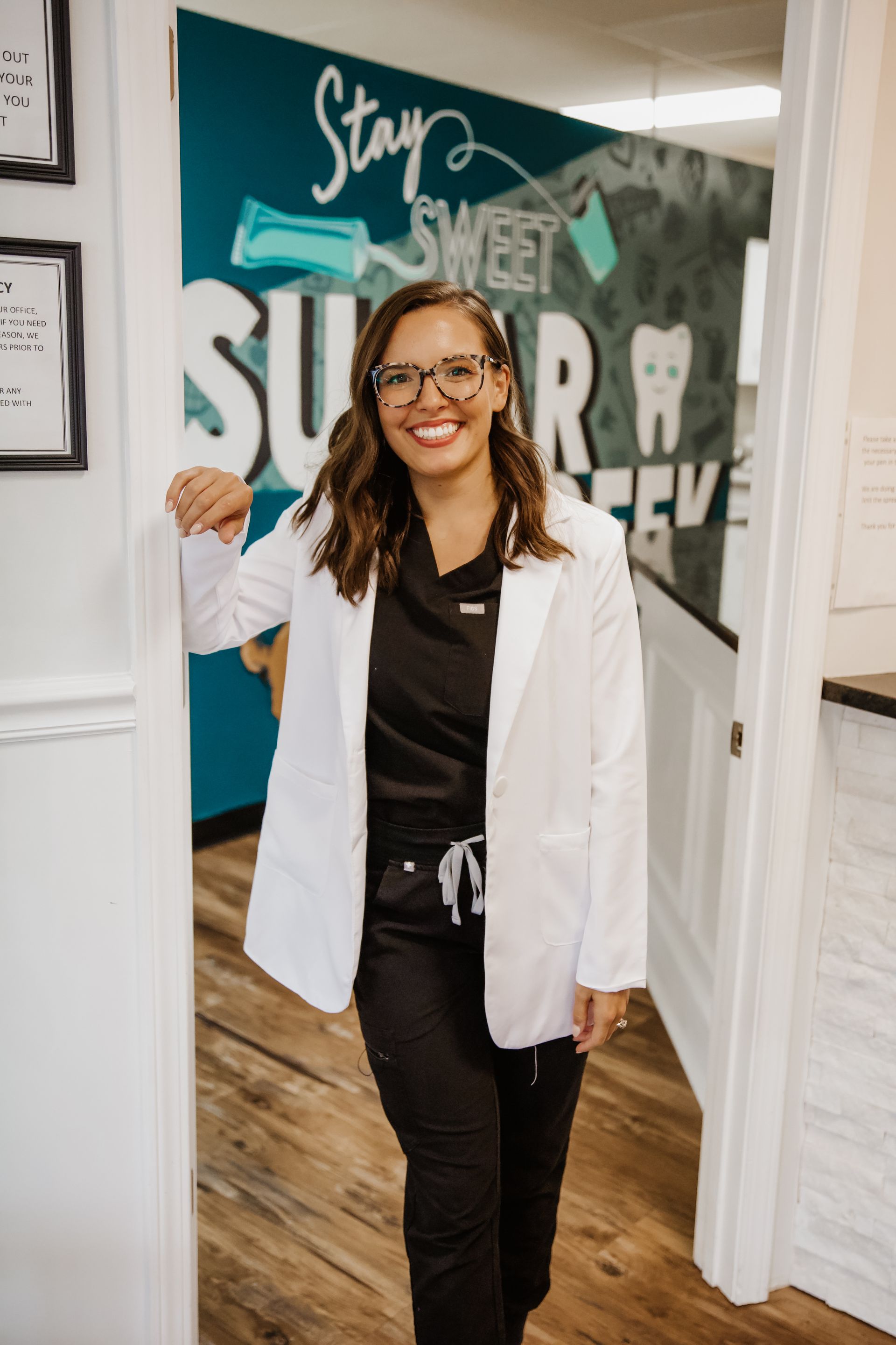 Smiling person in a white lab coat and scrubs, standing in a doorway of a dental office. 