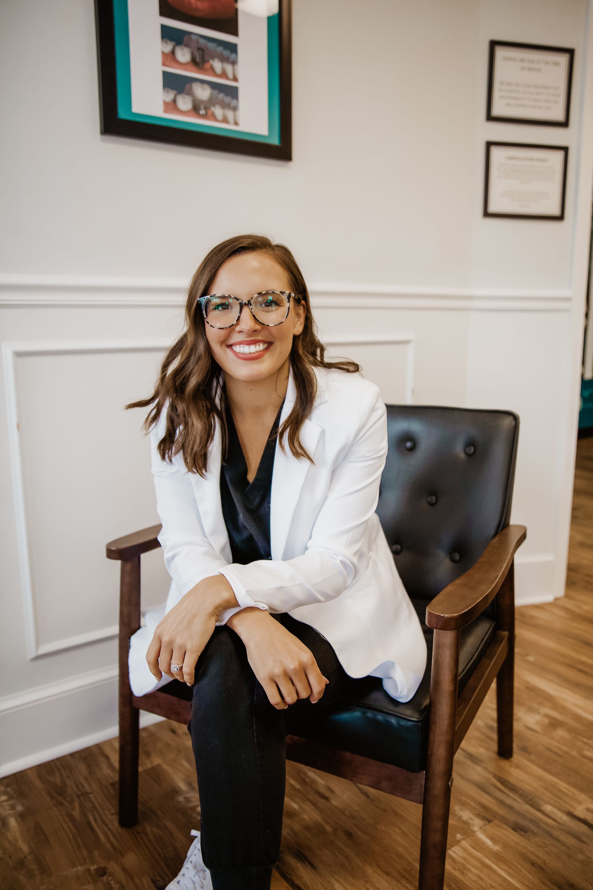 Woman in glasses and white blazer smiles, seated in a chair, wood-paneled room.