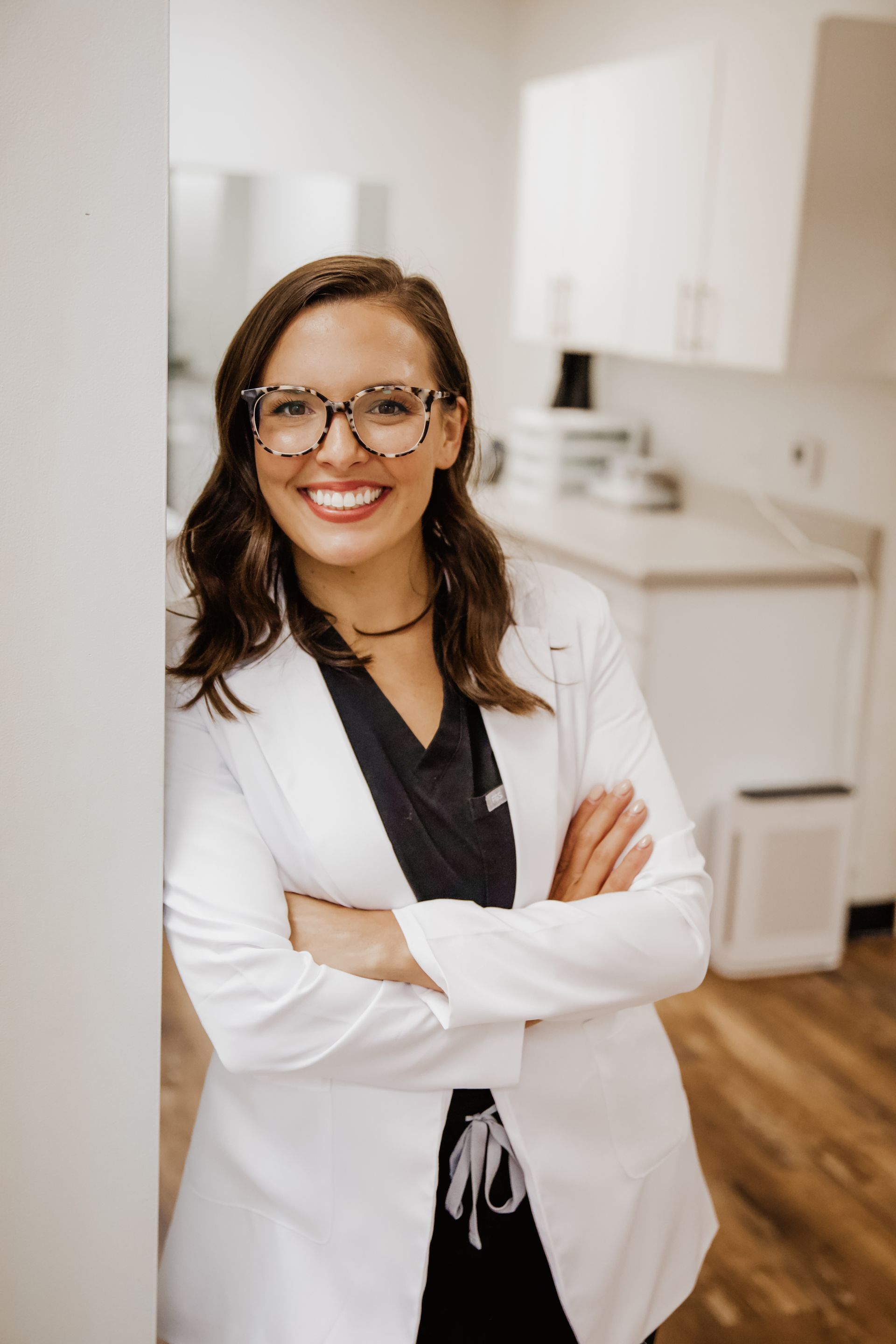 Woman in white coat, glasses, smiling with arms crossed in medical office.