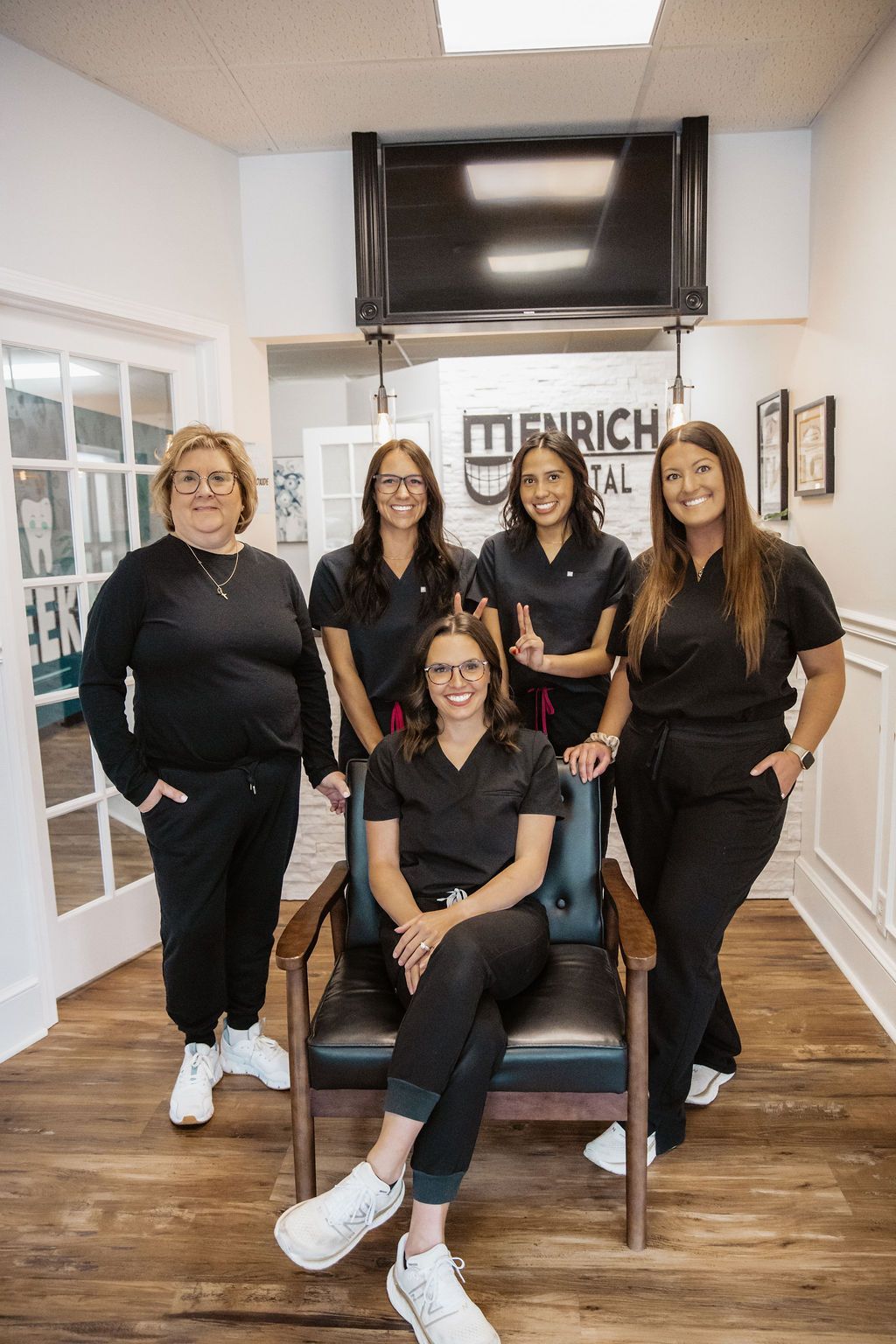 Five dental staff in black scrubs pose in an office. One sits in a chair, others stand, smiling.