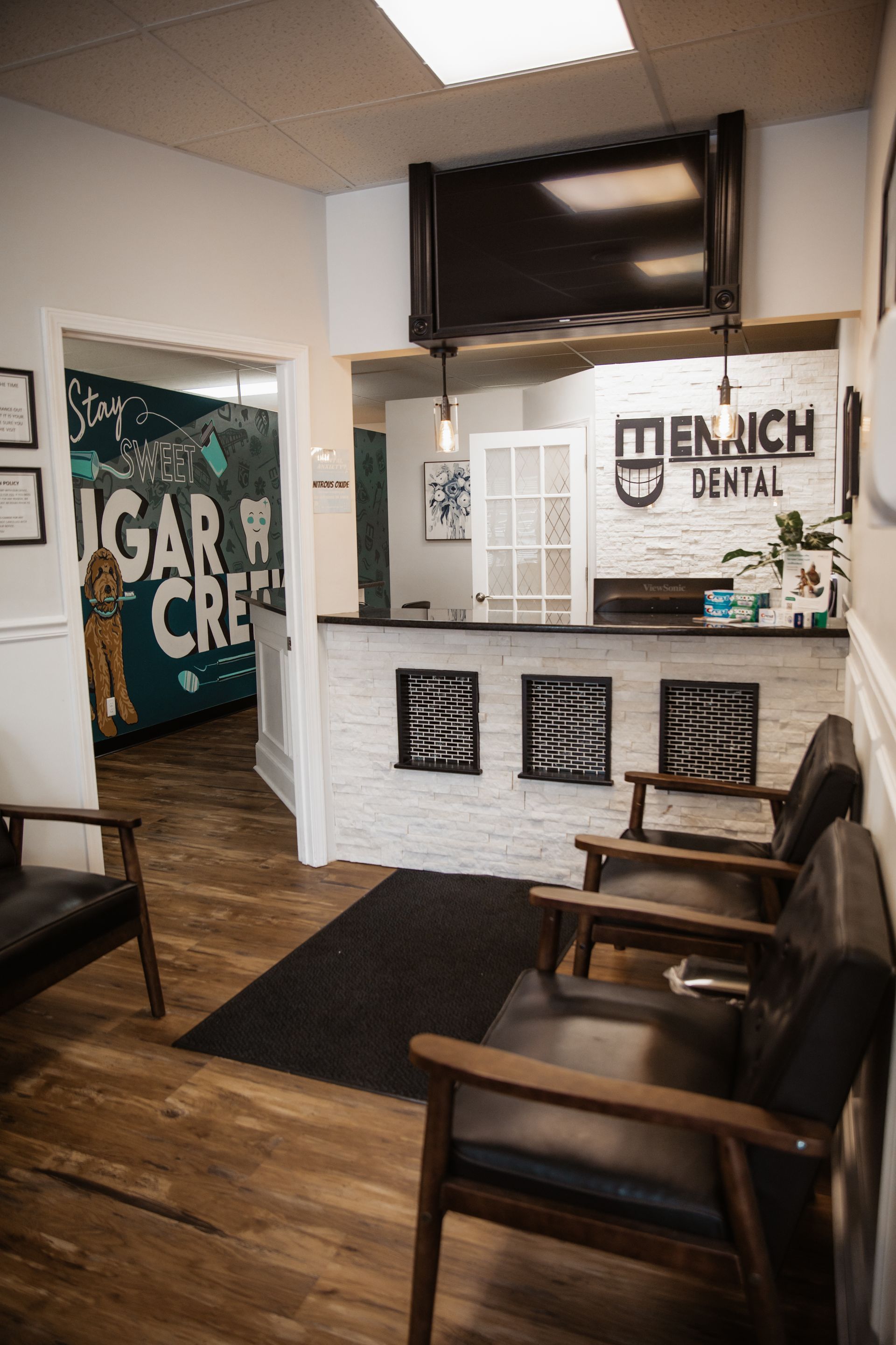 Waiting room with reception desk: chairs, brick wall, Sugar Creek mural, and a mounted TV.