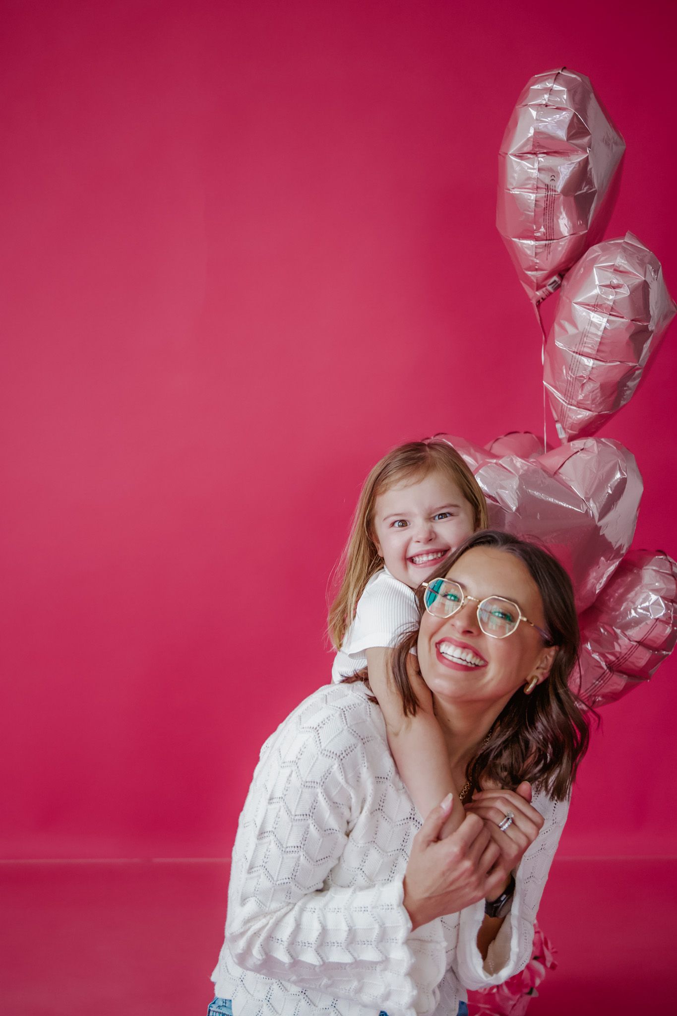 Woman with glasses, smiles while holding a child on her shoulders, pink balloons.