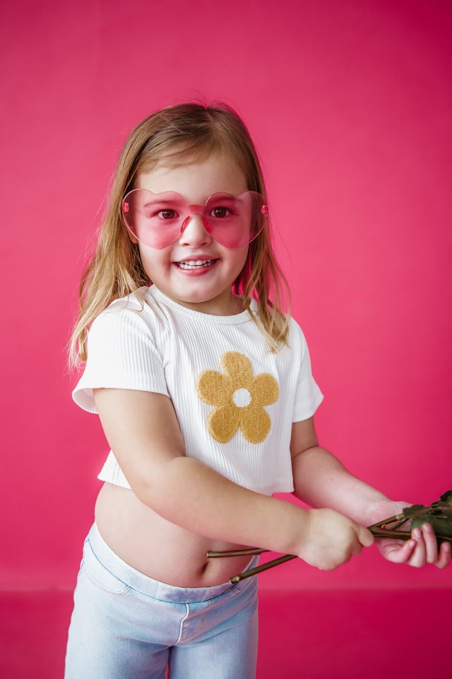 Girl with pink sunglasses and white shirt with daisy design, smiling in front of a pink background.