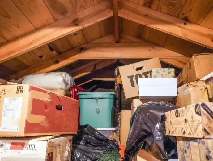 Cluttered attic storage with boxes, containers, and bags under a wooden ceiling.