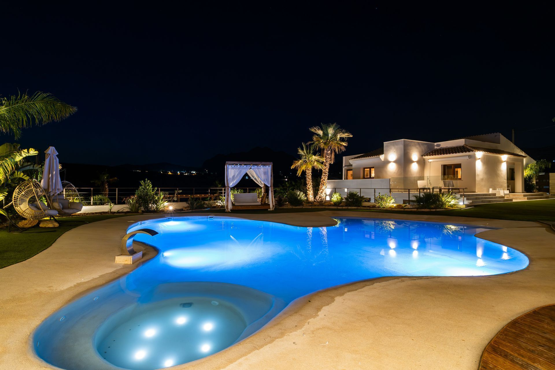 Night view of a lit pool next to a house with palm trees, a gazebo, and dark sky.