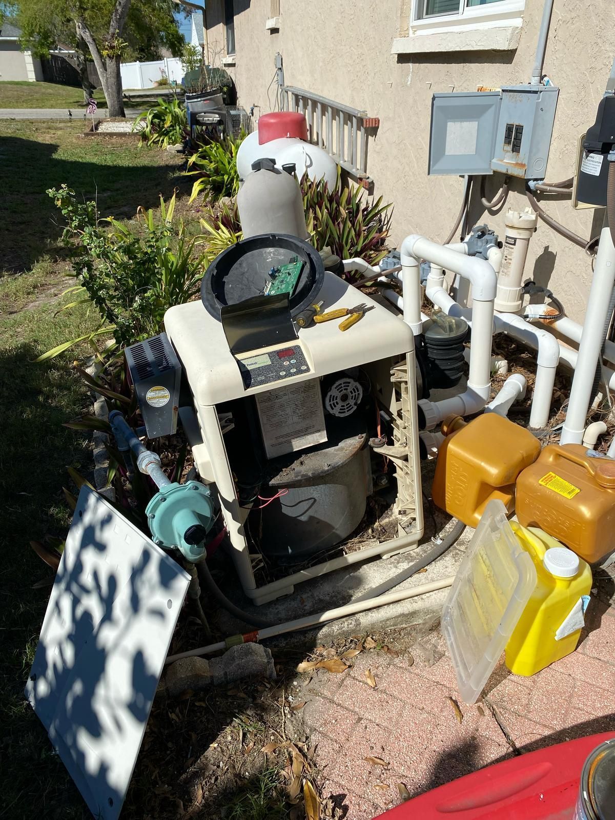 A pool heater with its cover removed, surrounded by piping and containers, sits near a house.