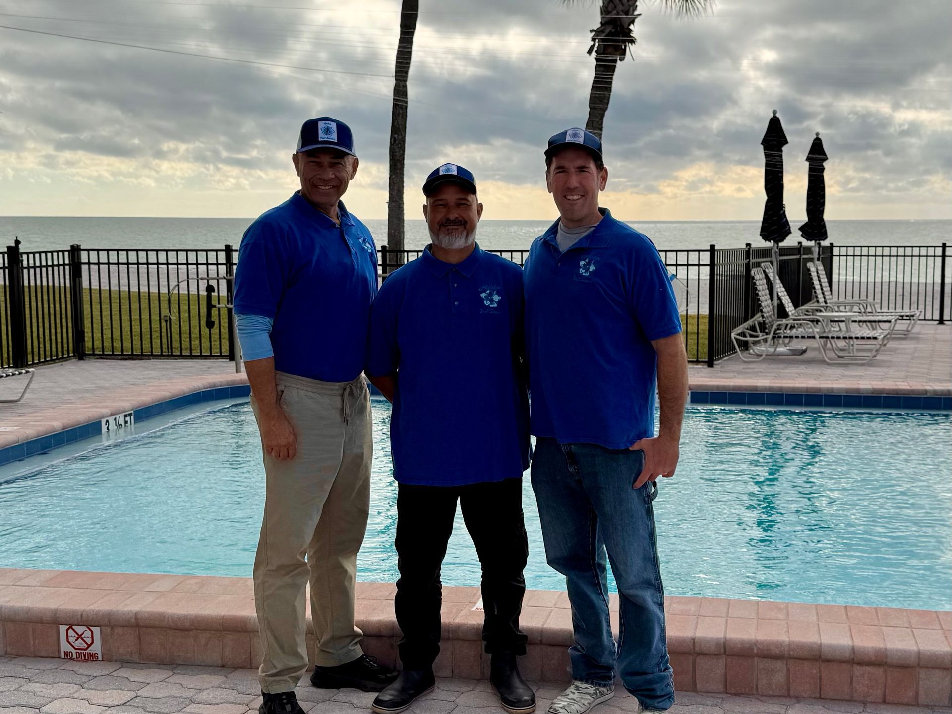 Three people wearing blue shirts and hats stand by a pool with the ocean in the background.