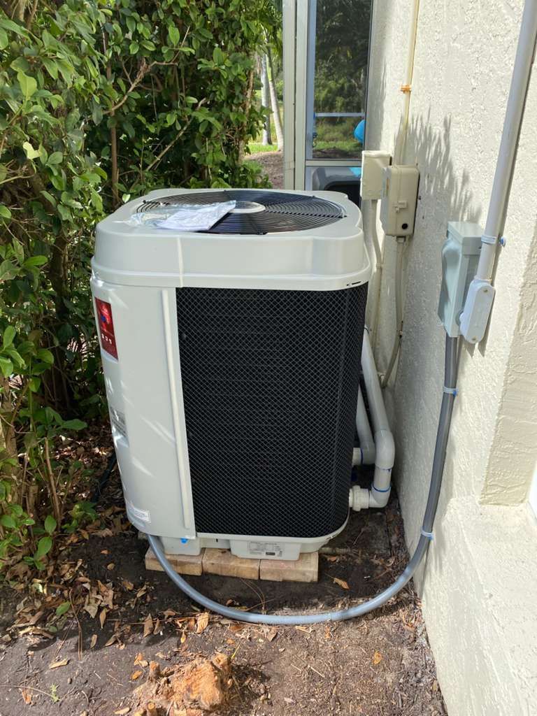 White pool heat pump next to a light-colored wall with gray conduit. Black mesh front, green foliage in the background.