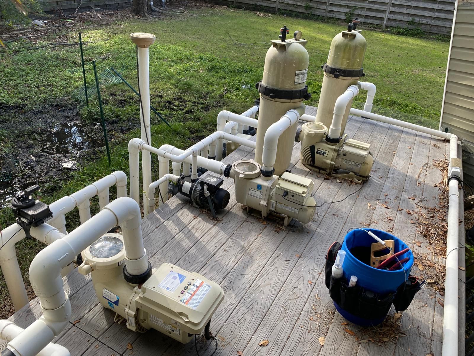 Pool pump equipment on a wooden deck, including pumps, filters, and plumbing. A blue bucket contains tools.