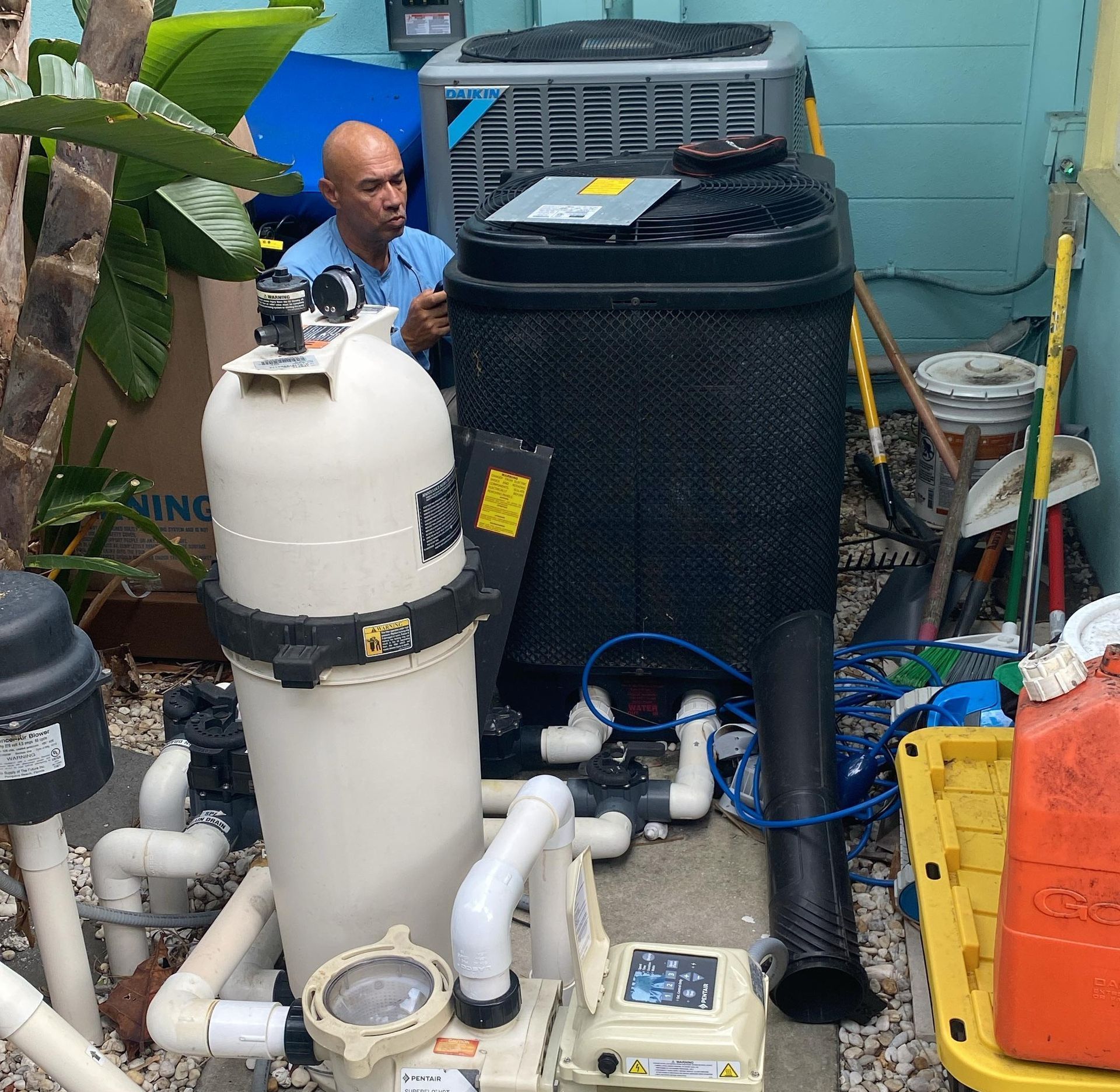 A man works on pool equipment. White filter, black heater, and pump in a small outdoor space.