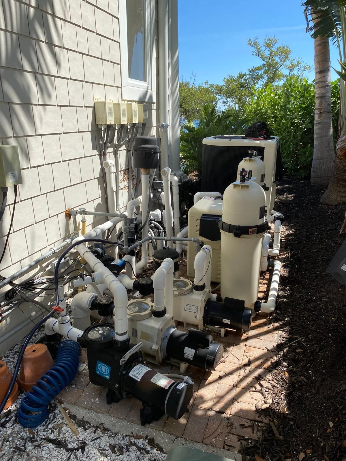 Outdoor pool equipment: pipes, pumps, and filters beside a house wall on a brick patio.