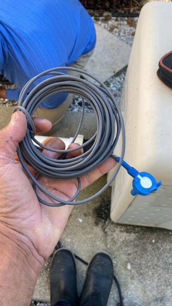 Hand holding a coiled gray cable with a blue connector outdoors near a vehicle and black boots